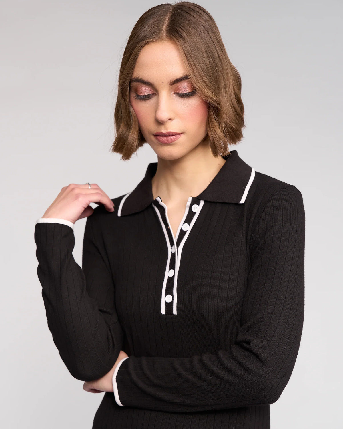 A woman with short, light brown hair wears the Ritual Dress in black by Knewe Label, featuring white trim and buttons, as she looks down with her left hand raised near her shoulder against a plain background.