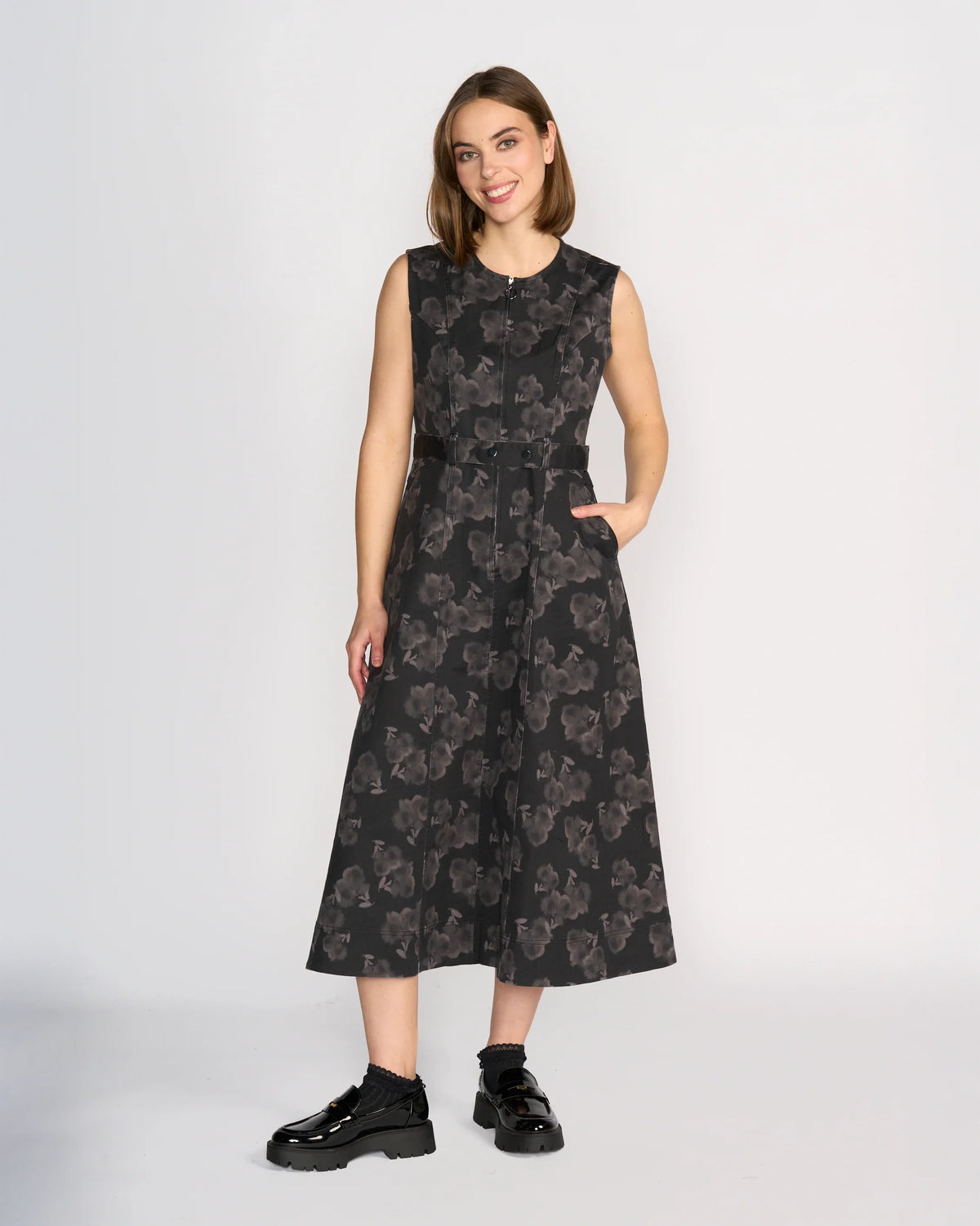 A woman with straight brown hair smiles against a plain white background, wearing the sleeveless Yuki Dress - Echo by Knewe Label— a dark midi dress with grey floral pattern and pockets—paired with black chunky shoes.