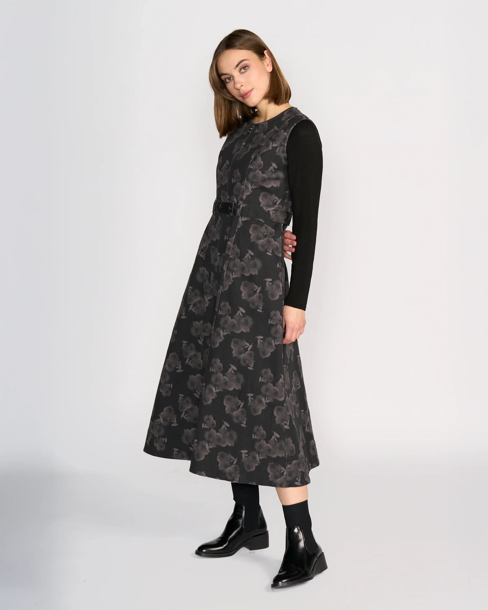 A woman with straight, shoulder-length hair stands against a plain background wearing the Knewe Label Yuki Dress - Echo over a long-sleeve black top and black ankle boots, looking slightly to the side.
