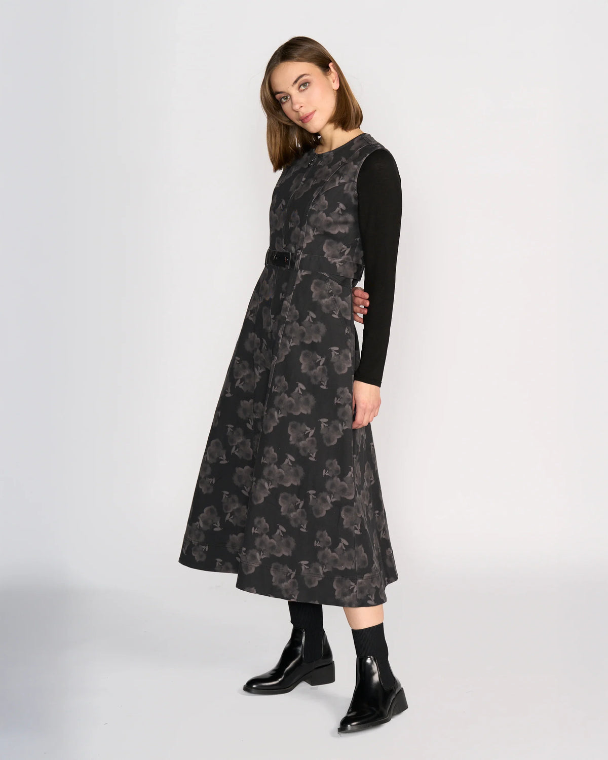 A woman with straight, shoulder-length hair stands against a plain background wearing the Knewe Label Yuki Dress - Echo over a long-sleeve black top and black ankle boots, looking slightly to the side.