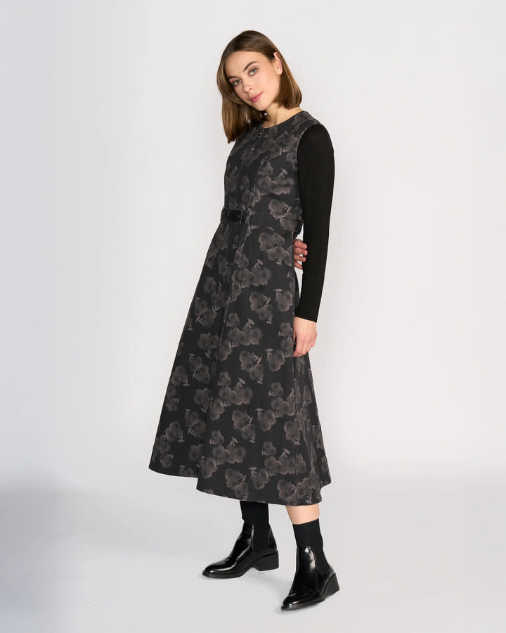 A woman with straight, shoulder-length hair stands against a plain background wearing the Knewe Label Yuki Dress - Echo over a long-sleeve black top and black ankle boots, looking slightly to the side.