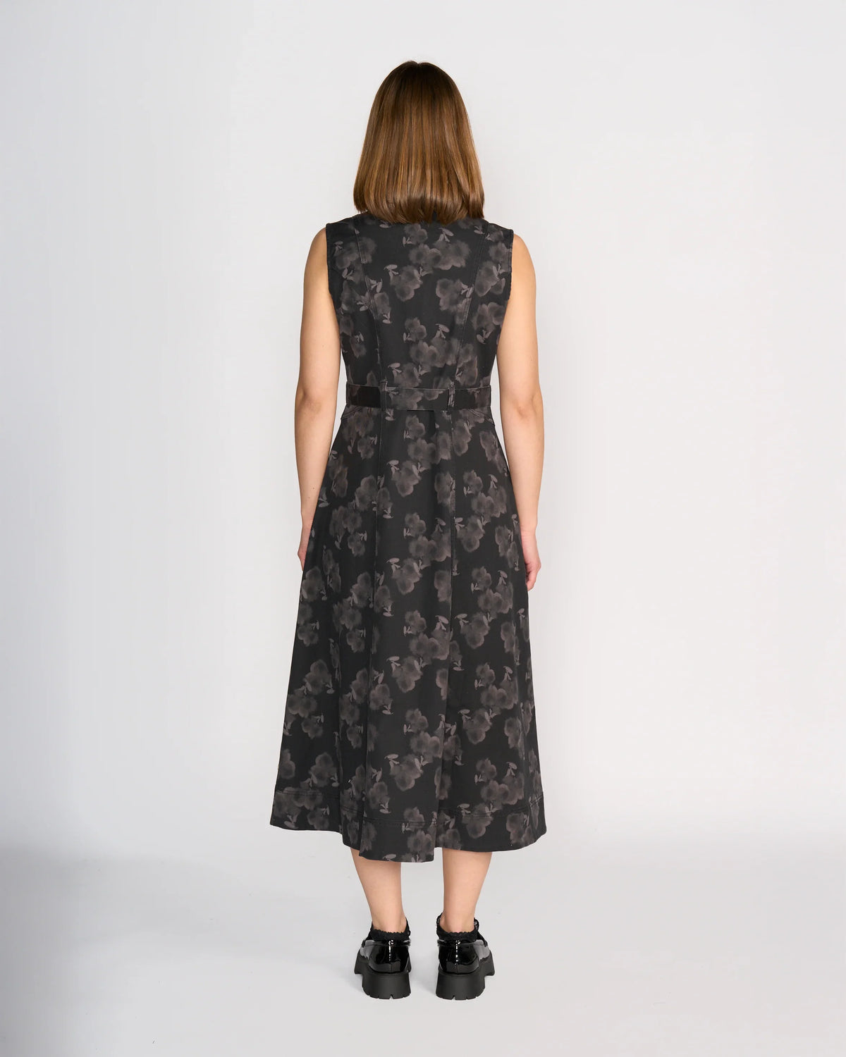 A woman with straight, shoulder-length brown hair faces away from the camera, wearing the Knewe Label Yuki Dress – Echo and black platform shoes. The sleeveless dress features a grey floral design. The background is plain white.