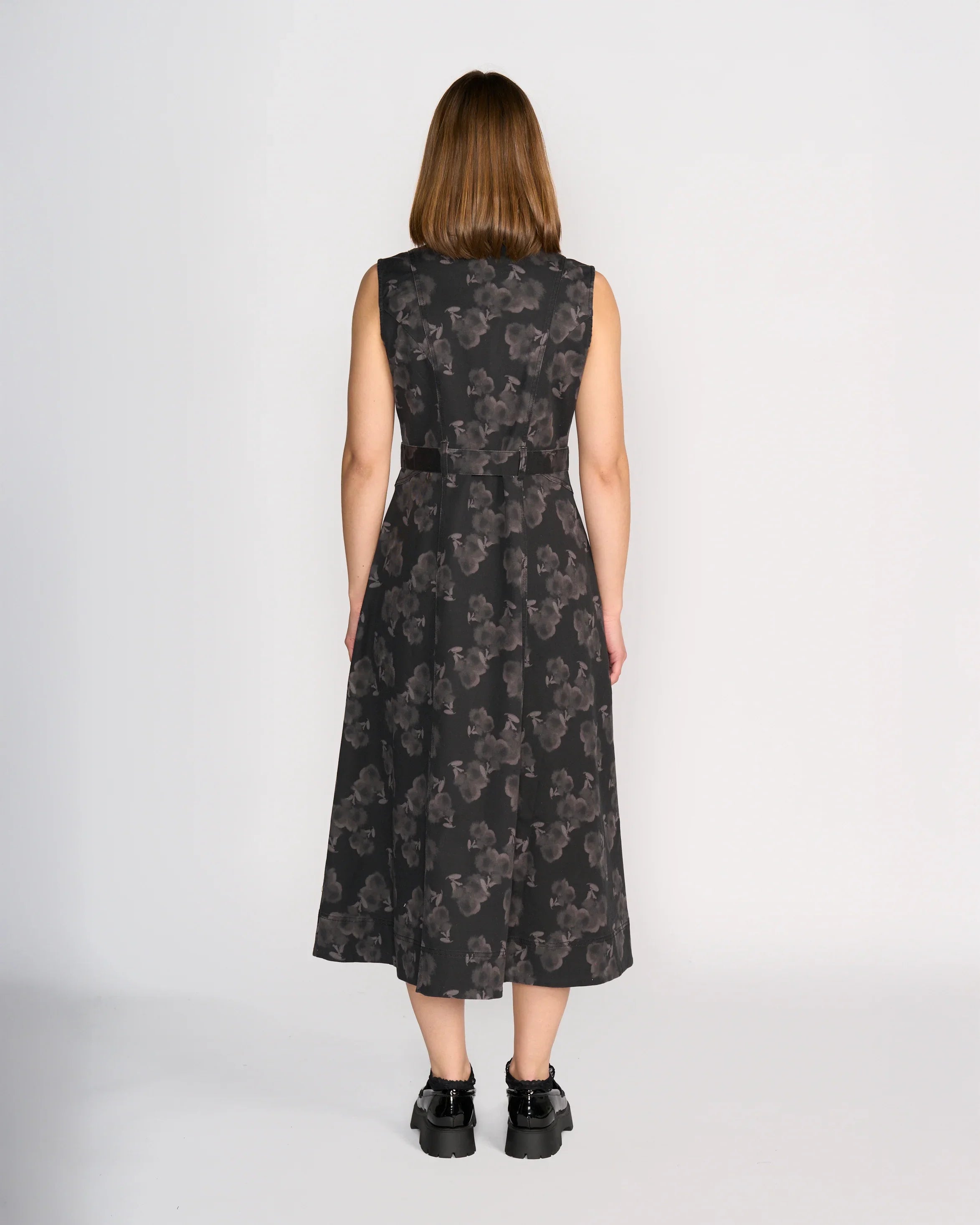 A woman with straight, shoulder-length brown hair faces away from the camera, wearing the Knewe Label Yuki Dress – Echo and black platform shoes. The sleeveless dress features a grey floral design. The background is plain white.