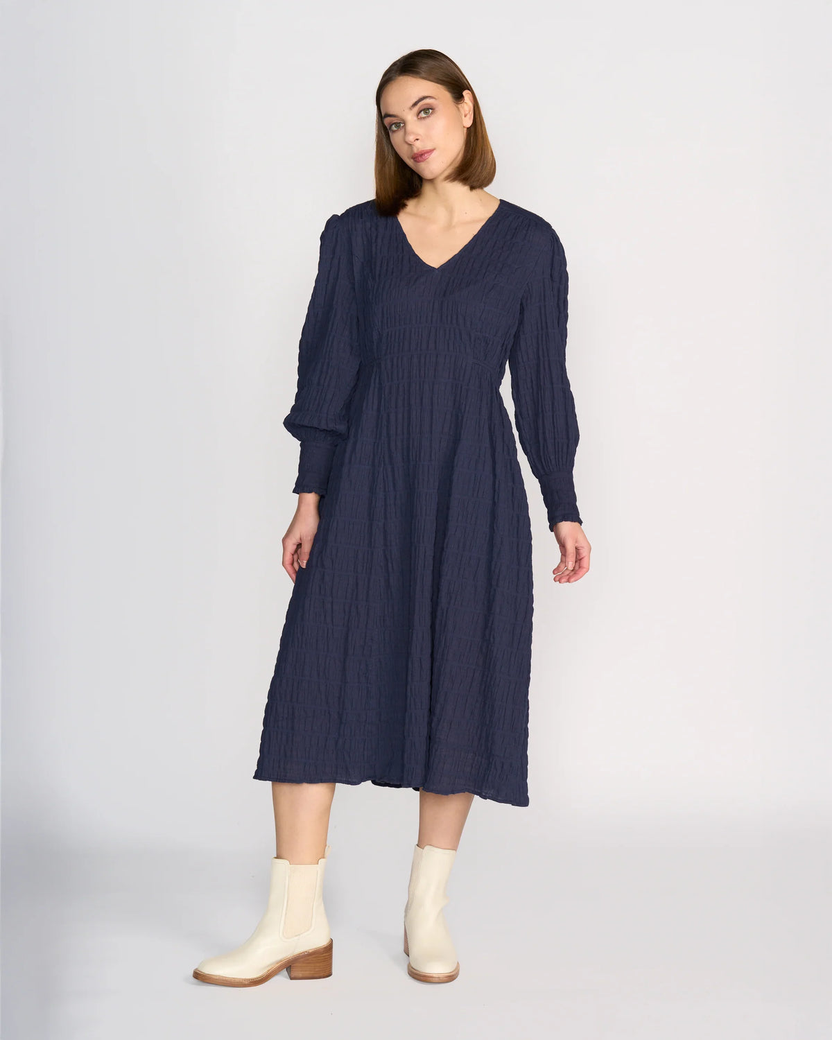 A woman wearing the Knewe Label Fleur Dress in Ink, a long sleeve winter dress with a V-neckline, and cream ankle boots stands against a plain light background.