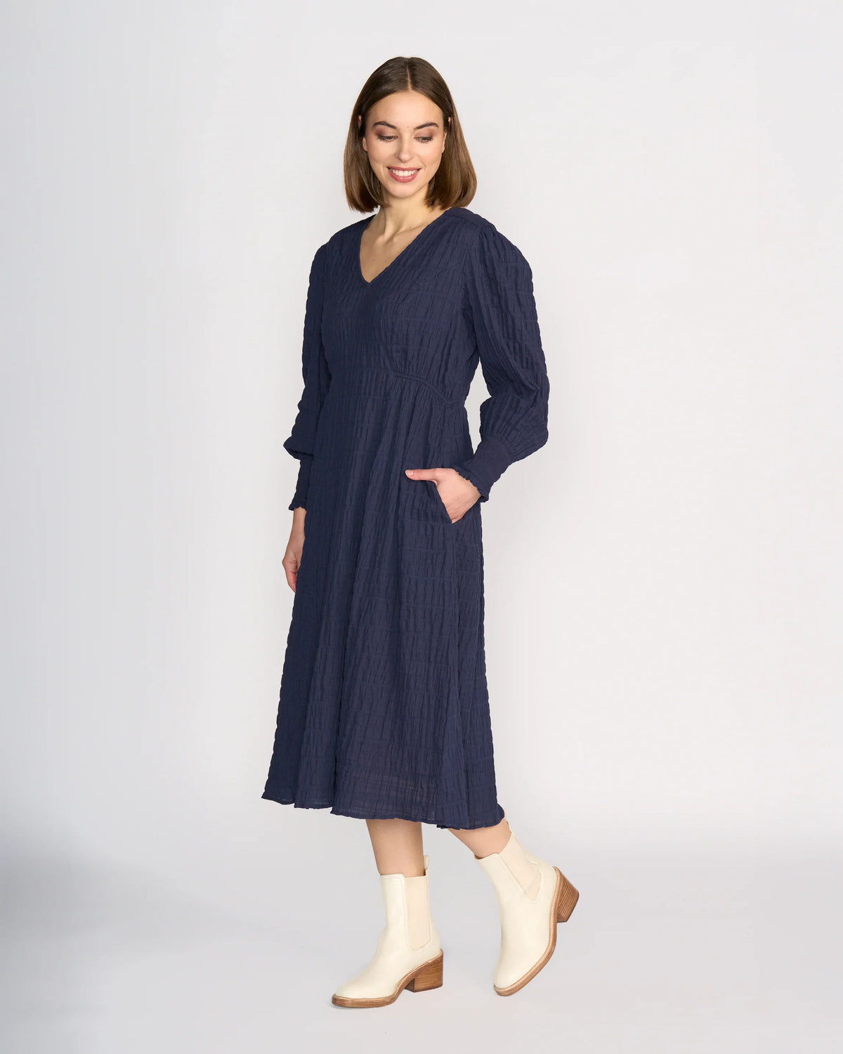 A woman models the Knewe Label Fleur Dress - Ink, a long sleeve navy midi with a V-neckline and side pockets, against a plain white background. She looks down, smiling, and pairs it with cream-colored ankle boots.