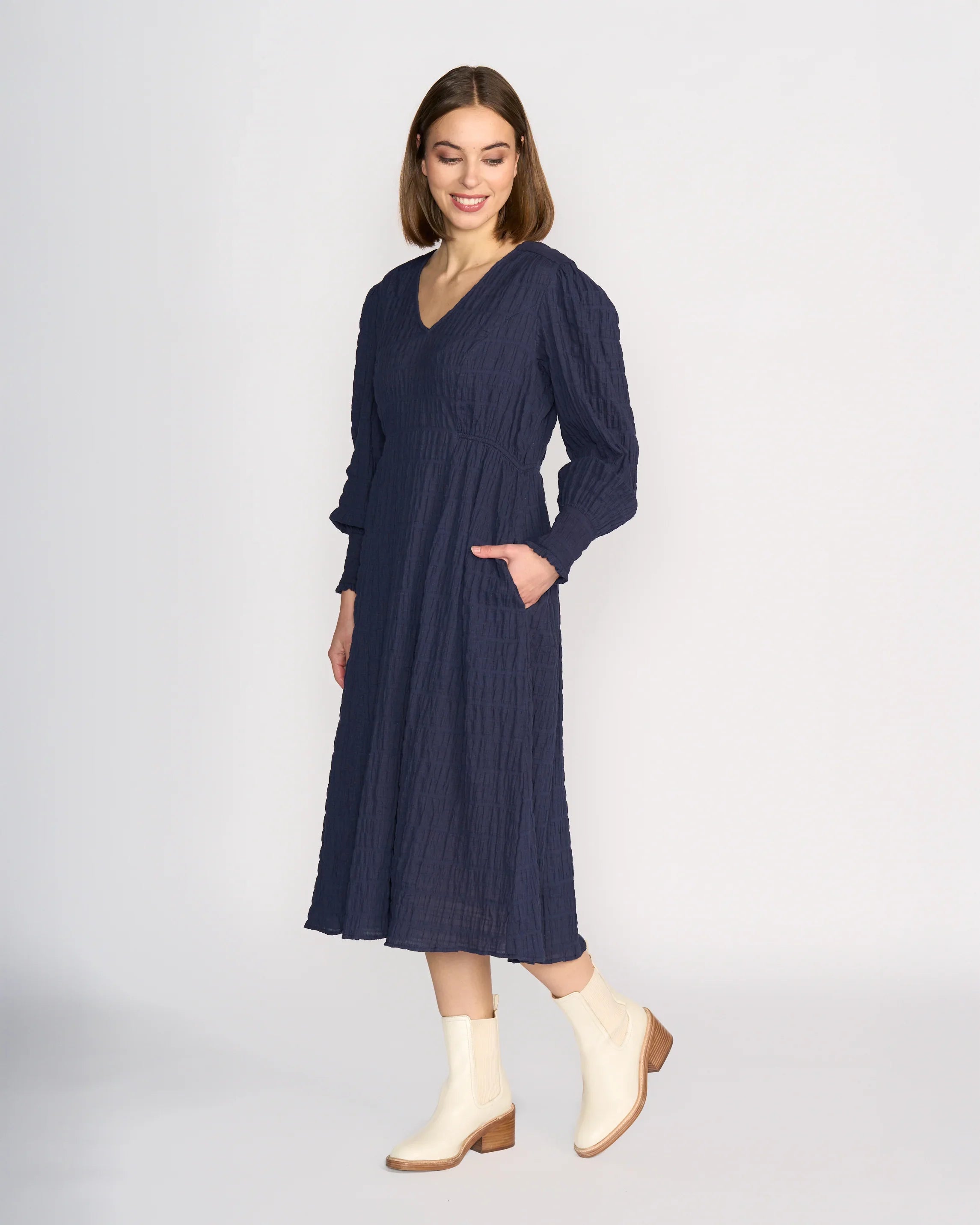 A woman models the Knewe Label Fleur Dress - Ink, a long sleeve navy midi with a V-neckline and side pockets, against a plain white background. She looks down, smiling, and pairs it with cream-colored ankle boots.