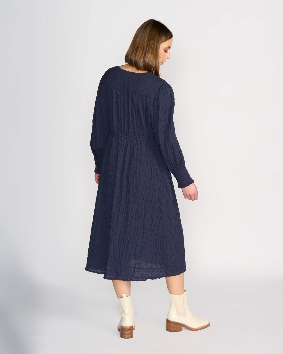 A woman faces away from the camera in the Knewe Label Fleur Dress - Ink, a long sleeve navy winter dress with a V-neckline, paired with white ankle boots featuring brown heels, set against a plain light background.