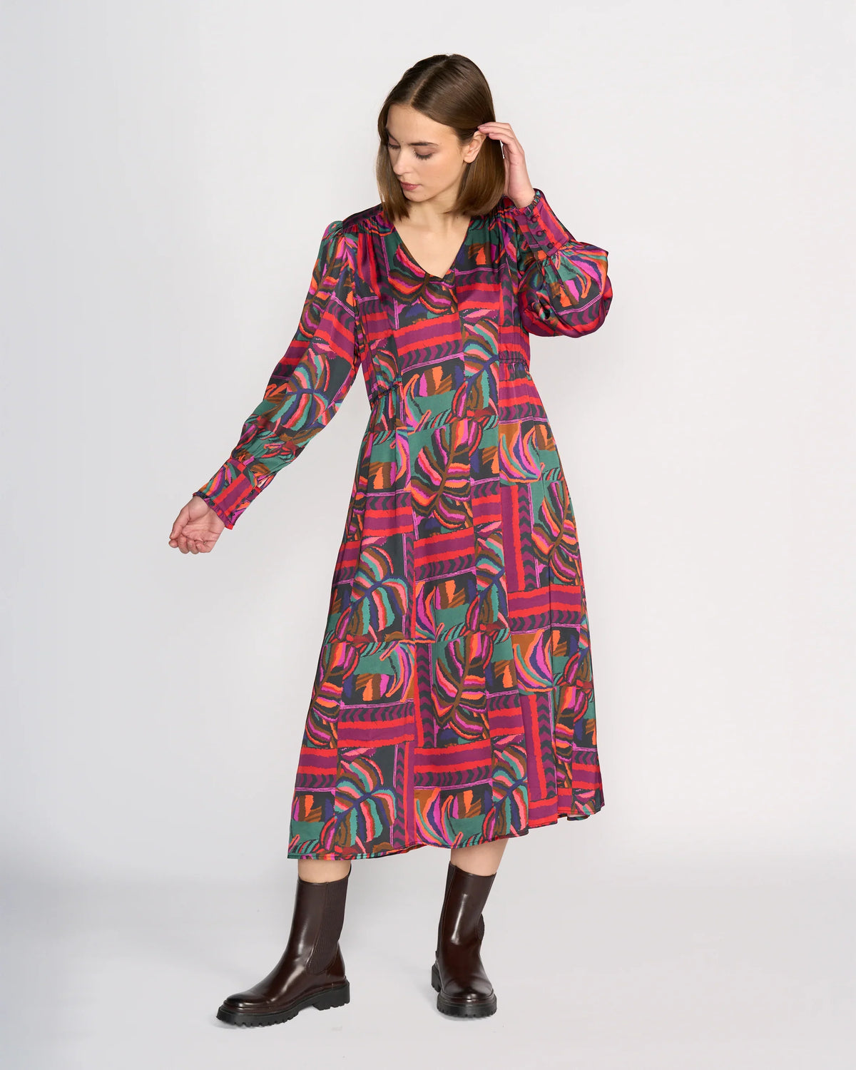 A woman models the Knewe Label Fleur Dress - Sunset, a colorful long-sleeved midi in bold red, green, and purple geometric and leaf patterns, paired with dark boots against a plain white background.