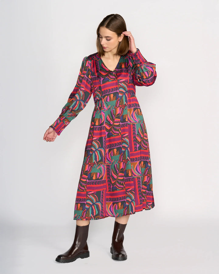 A woman models the Knewe Label Fleur Dress - Sunset, a colorful long-sleeved midi in bold red, green, and purple geometric and leaf patterns, paired with dark boots against a plain white background.