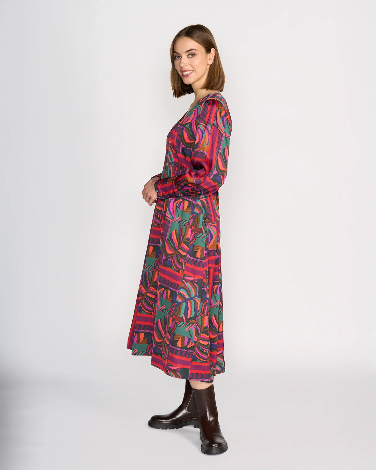 A woman stands sideways, smiling, wearing the Knewe Label Fleur Dress - Sunset in red, green, and purple tones with long sleeves and dark brown ankle boots, set against a plain white background.