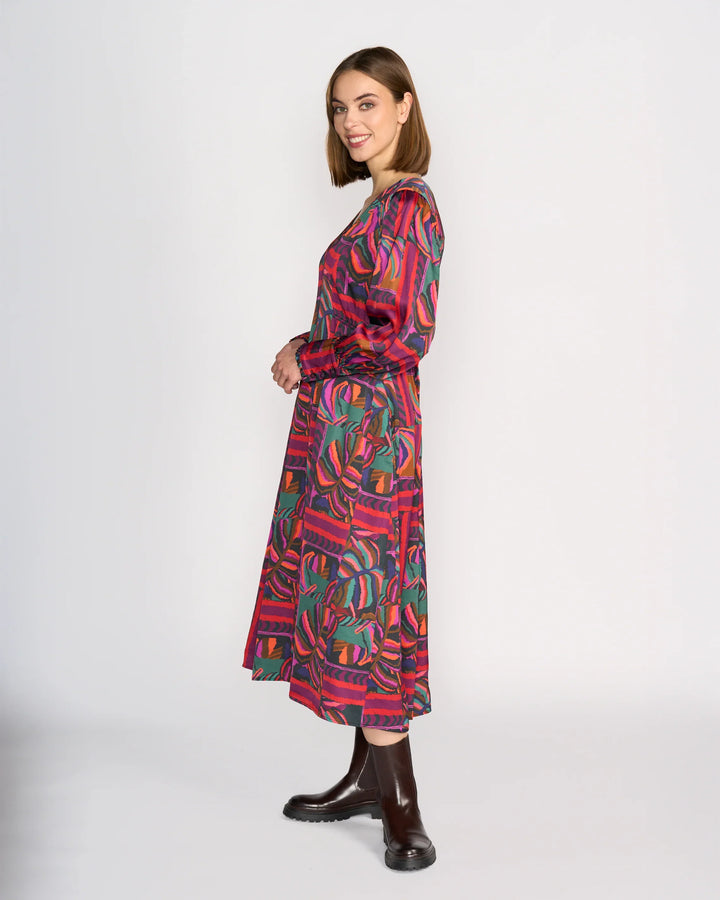 A woman stands sideways, smiling, wearing the Knewe Label Fleur Dress - Sunset in red, green, and purple tones with long sleeves and dark brown ankle boots, set against a plain white background.