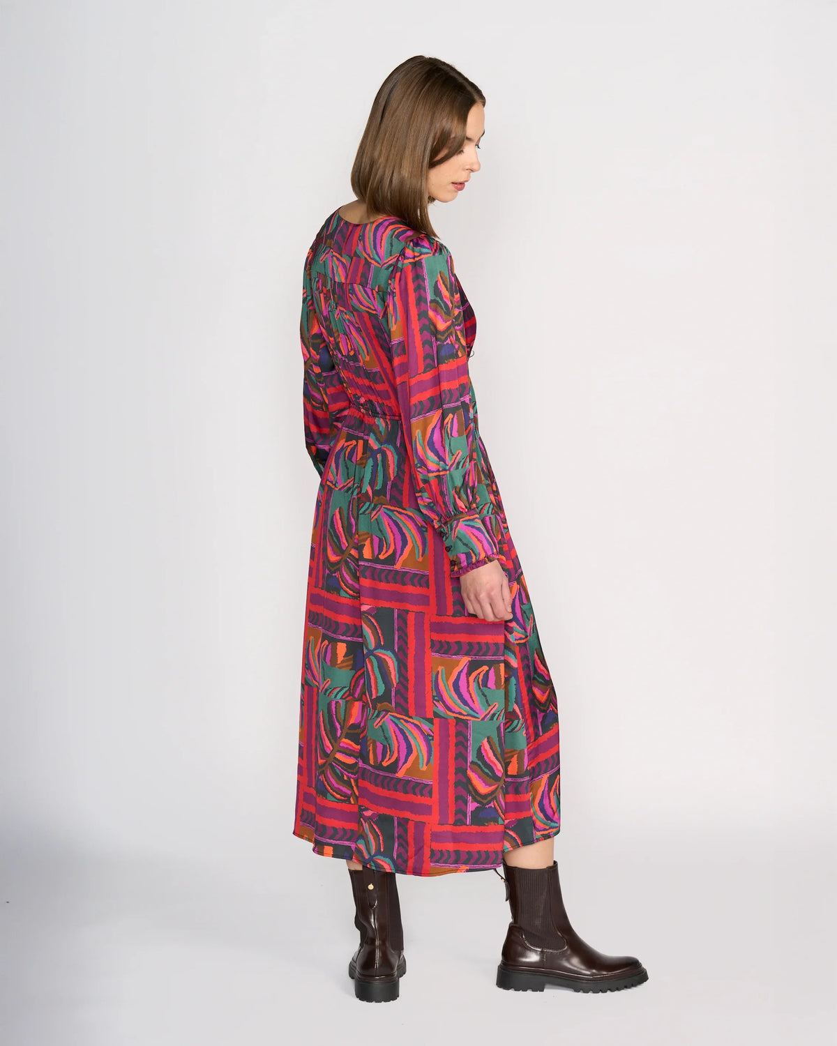 A woman with shoulder-length brown hair stands sideways wearing the Knewe Label Fleur Dress - Sunset, a long-sleeved, mid-length winter dress featuring bold green, purple, and pink geometric and leaf patterns. She pairs it with dark brown ankle boots.