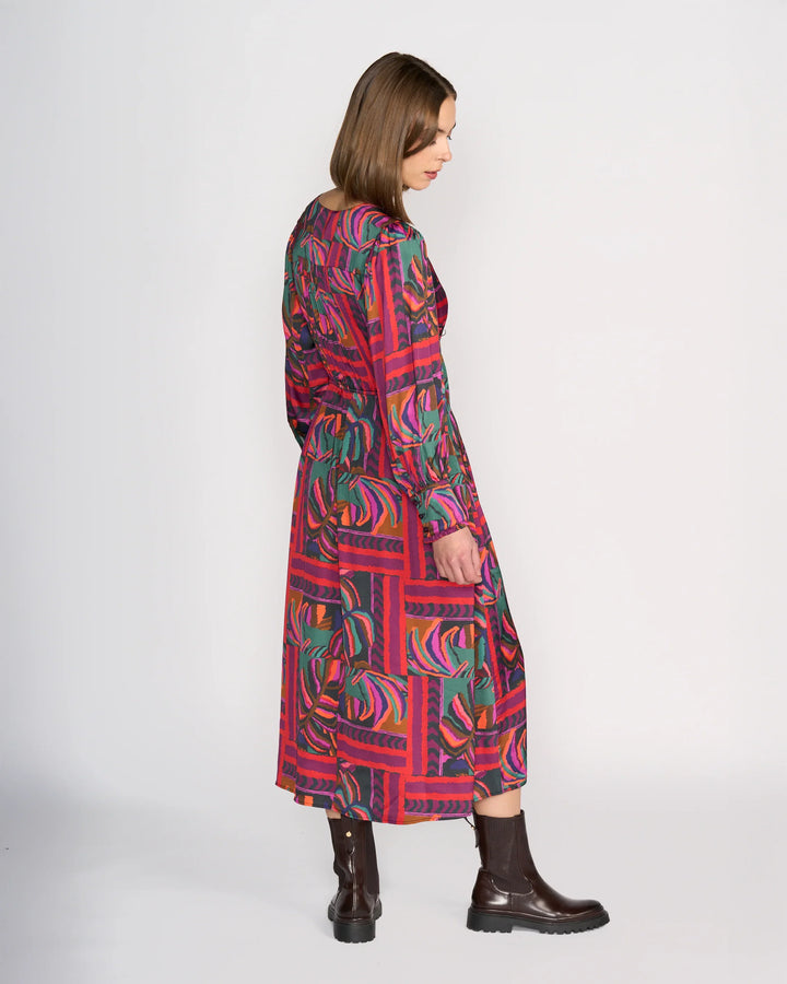 A woman with shoulder-length brown hair stands sideways wearing the Knewe Label Fleur Dress - Sunset, a long-sleeved, mid-length winter dress featuring bold green, purple, and pink geometric and leaf patterns. She pairs it with dark brown ankle boots.