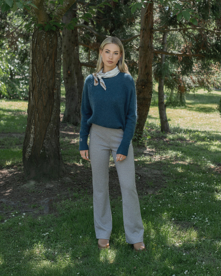 A woman stands on grass in a sunlit park, wearing Knewe Label’s Como Flare Pant in Cedar HT, paired with a blue sweater, brown shoes, and a neck scarf. Tall trees and dappled sunlight fill the background.
