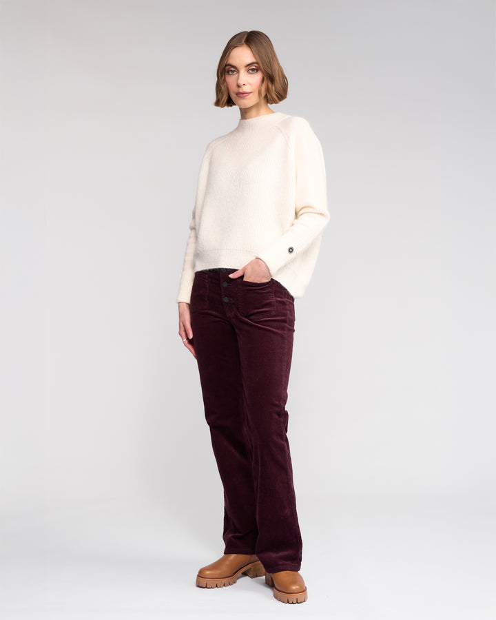 A woman stands against a plain background wearing the Knewe Label Mia Sweater in Winter White, paired with dark burgundy corduroy pants and tan chunky-soled shoes. One hand rests in her pocket as she gazes slightly to the side.