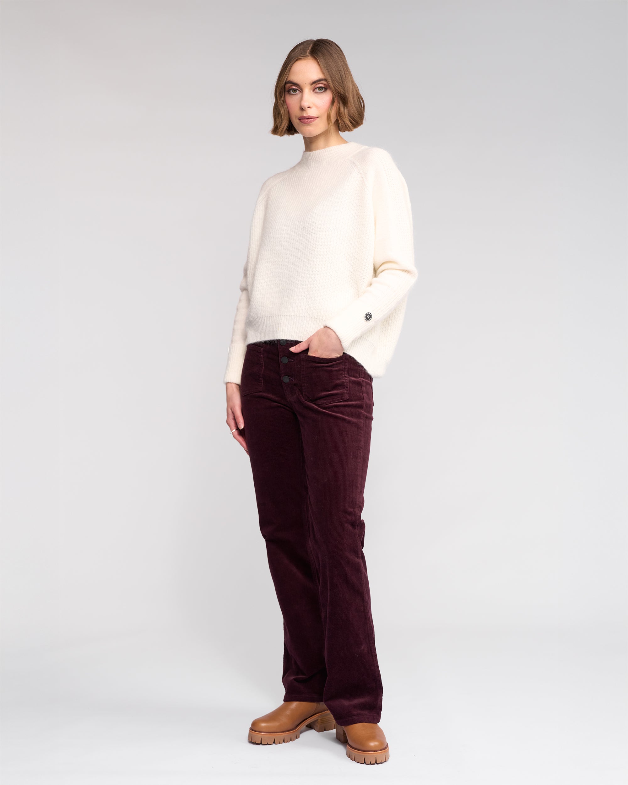 A woman stands against a plain background in the Knewe Label Mia Sweater in Winter White, paired with dark burgundy corduroy pants and tan platform shoes, one hand in her pocket and wearing a neutral expression.