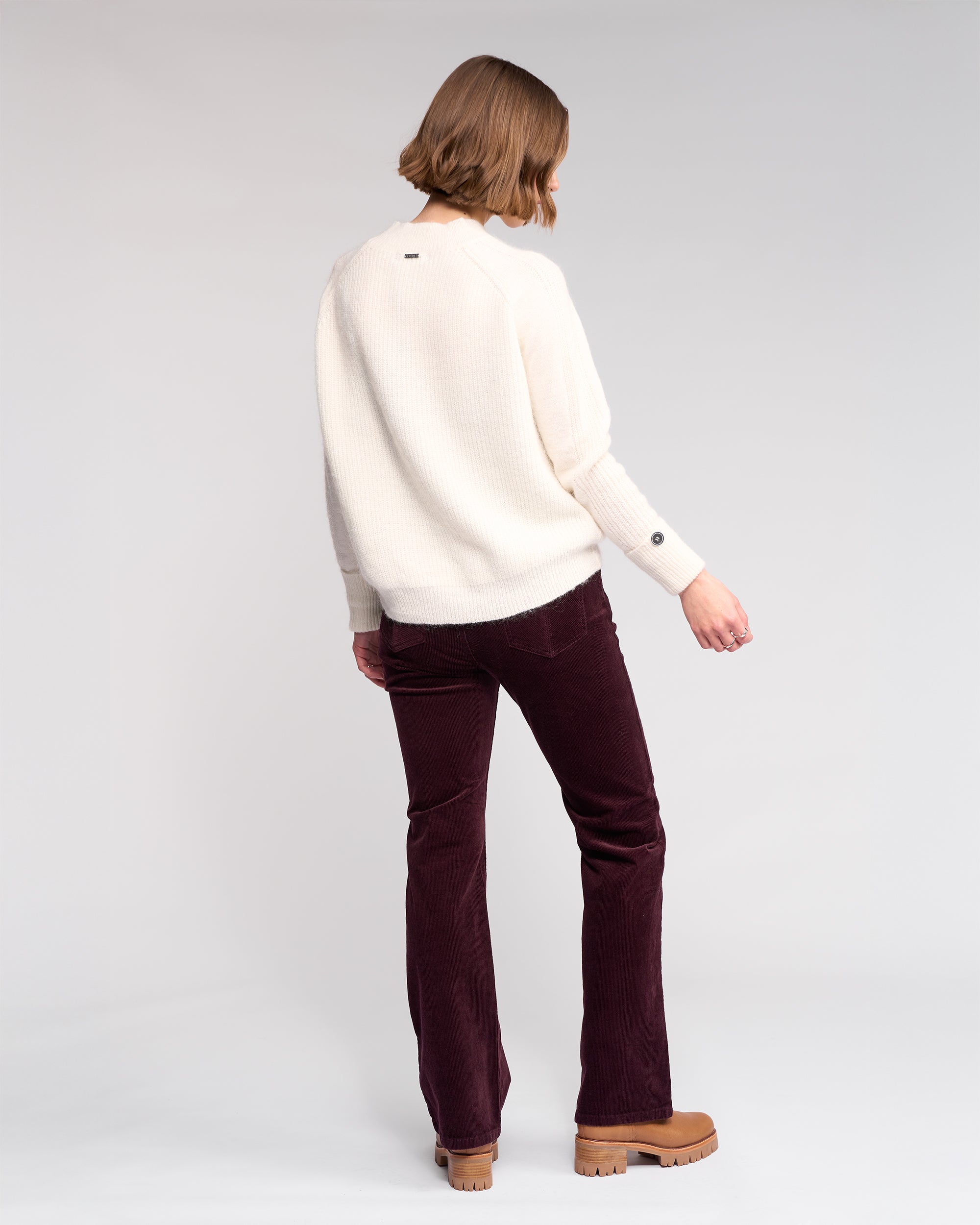 A person with short hair stands facing away, wearing the Knewe Label Mia Sweater in Winter White, dark purple corduroy pants, and tan platform shoes against a plain light background.