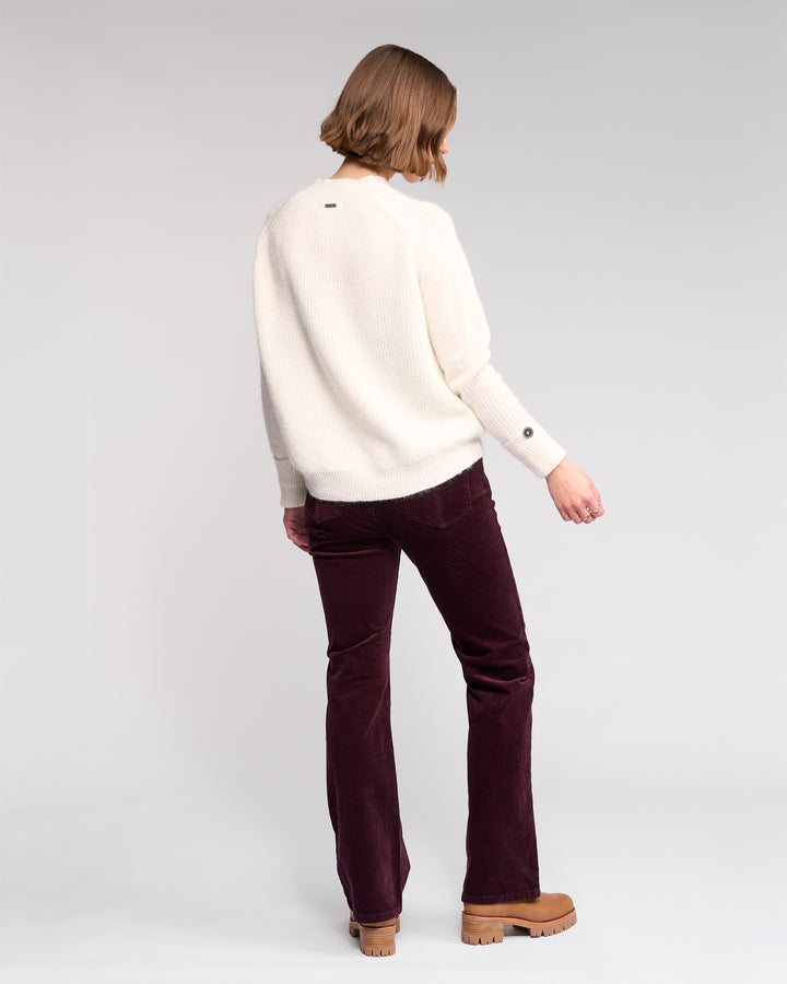 A person with short hair stands facing away, wearing the Knewe Label Mia Sweater in Winter White, dark purple corduroy pants, and tan platform shoes against a plain light background.