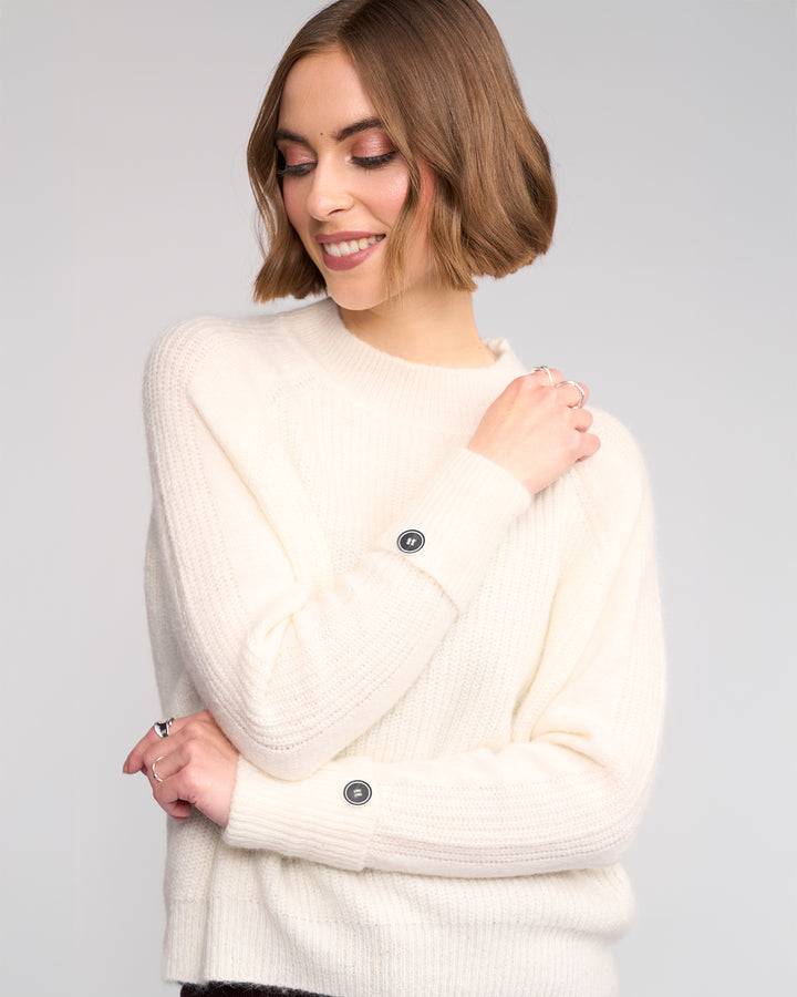 A woman with short, light brown hair is smiling and looking down while wearing the Knewe Label Mia Sweater in Winter White, a relaxed-fit knit jumper featuring decorative sleeve buttons and her arms crossed in front.