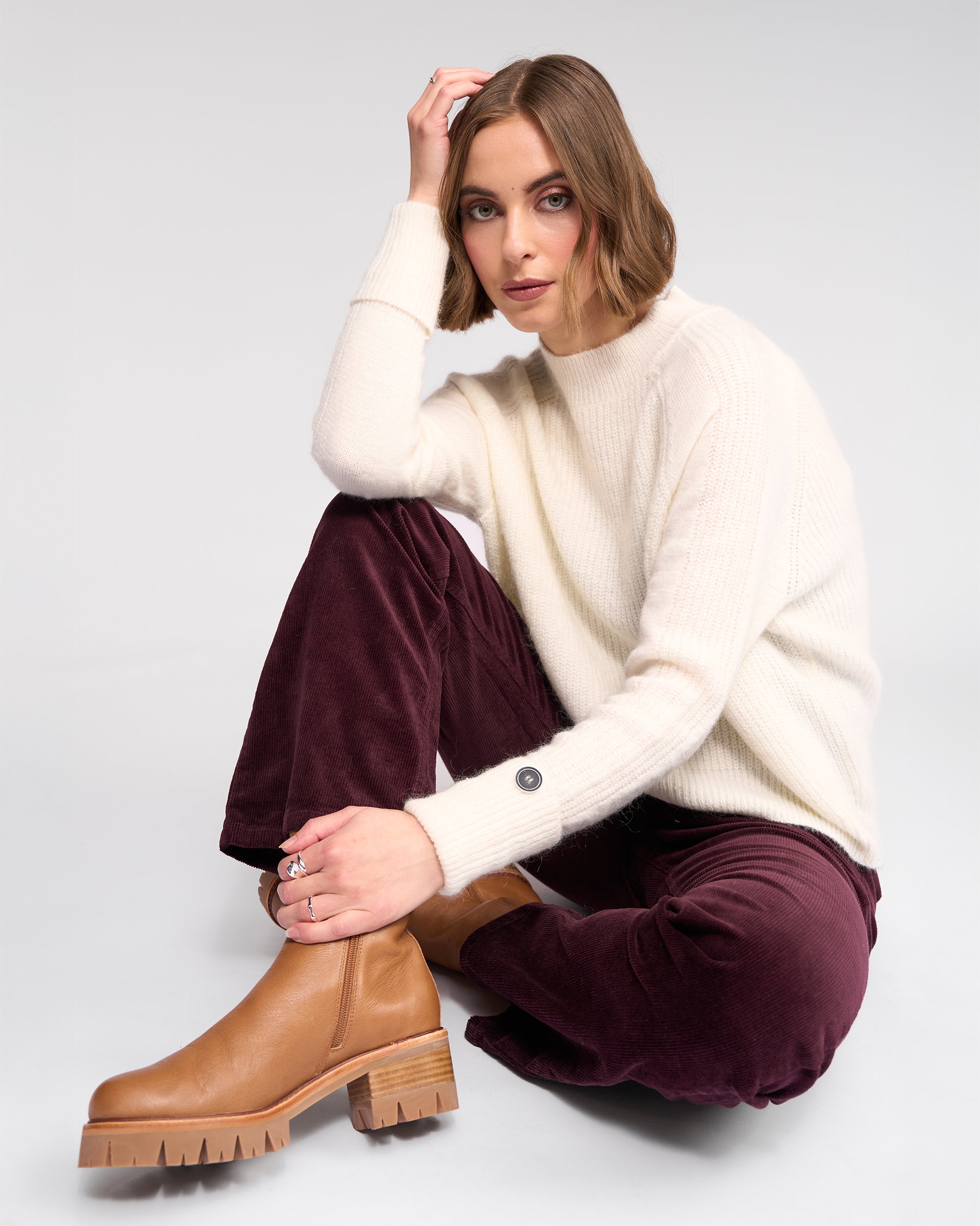 A woman with chin-length brown hair sits on the floor, wearing the Mia Sweater in Winter White by Knewe Label, paired with burgundy pants and tan chunky boots. She rests her elbow on her knee and looks at the camera neutrally.