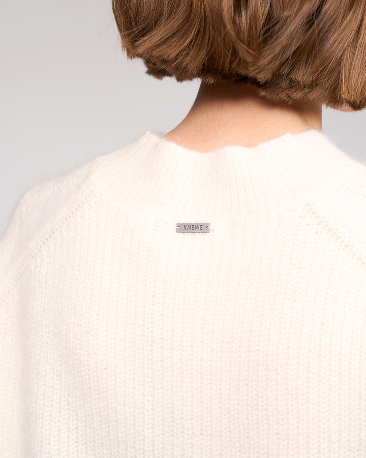 Close-up of the back of a person’s neck and shoulders wearing the Mia Sweater in Winter White by Knewe Label, a relaxed-fit knit. The short brown hair sits above the collar, with a small "KNEWE" tag at the back neckline.