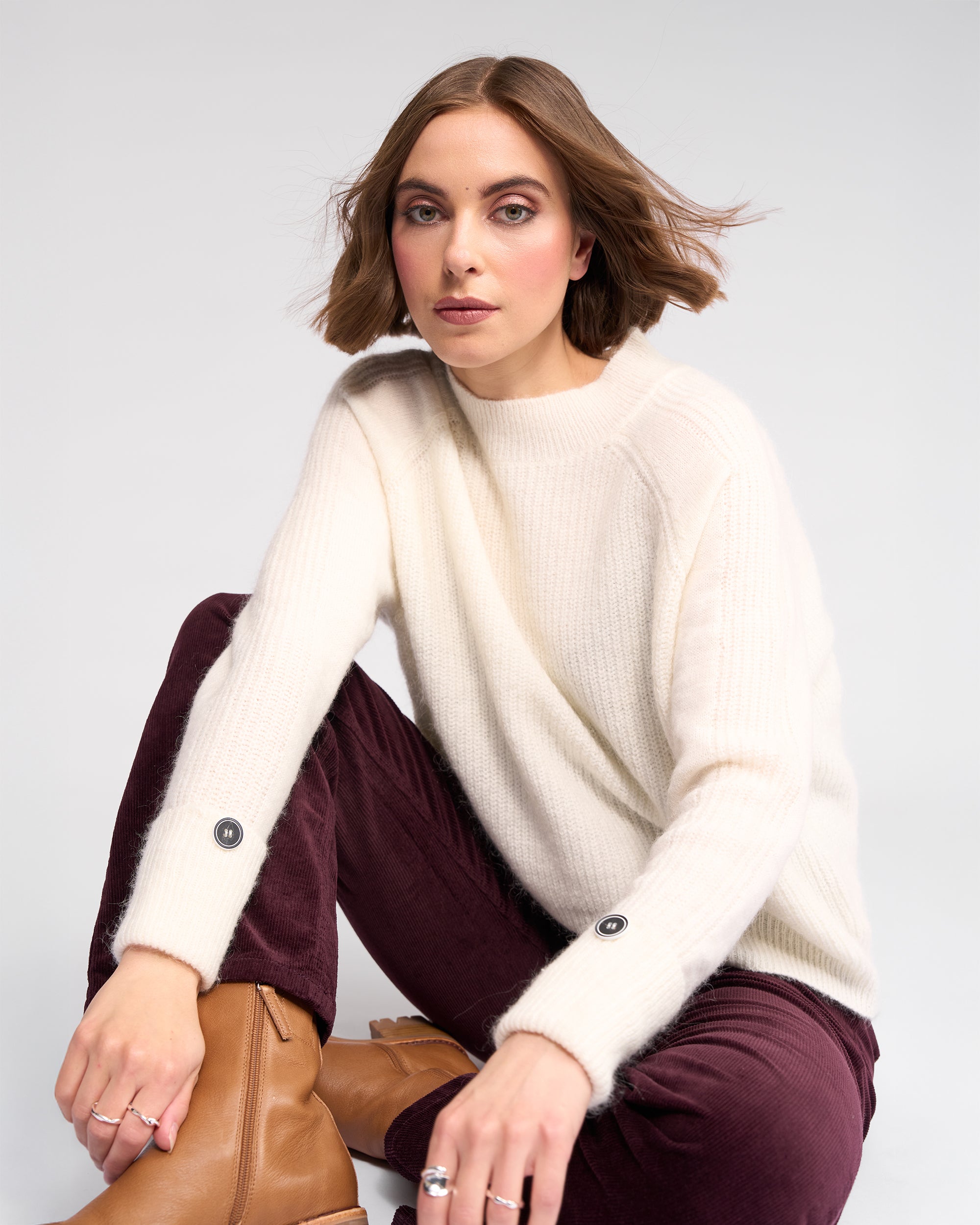 A woman with light brown bobbed hair wears the Mia Sweater in Winter White by Knewe Label, paired with burgundy pants and tan boots, sitting with one knee up against a plain light background.