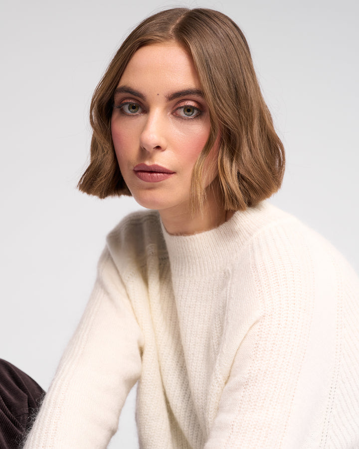 A woman with chin-length light brown hair and blue eyes wears the Knewe Label Mia Sweater in Winter White, gazing neutrally at the camera against a plain light background.