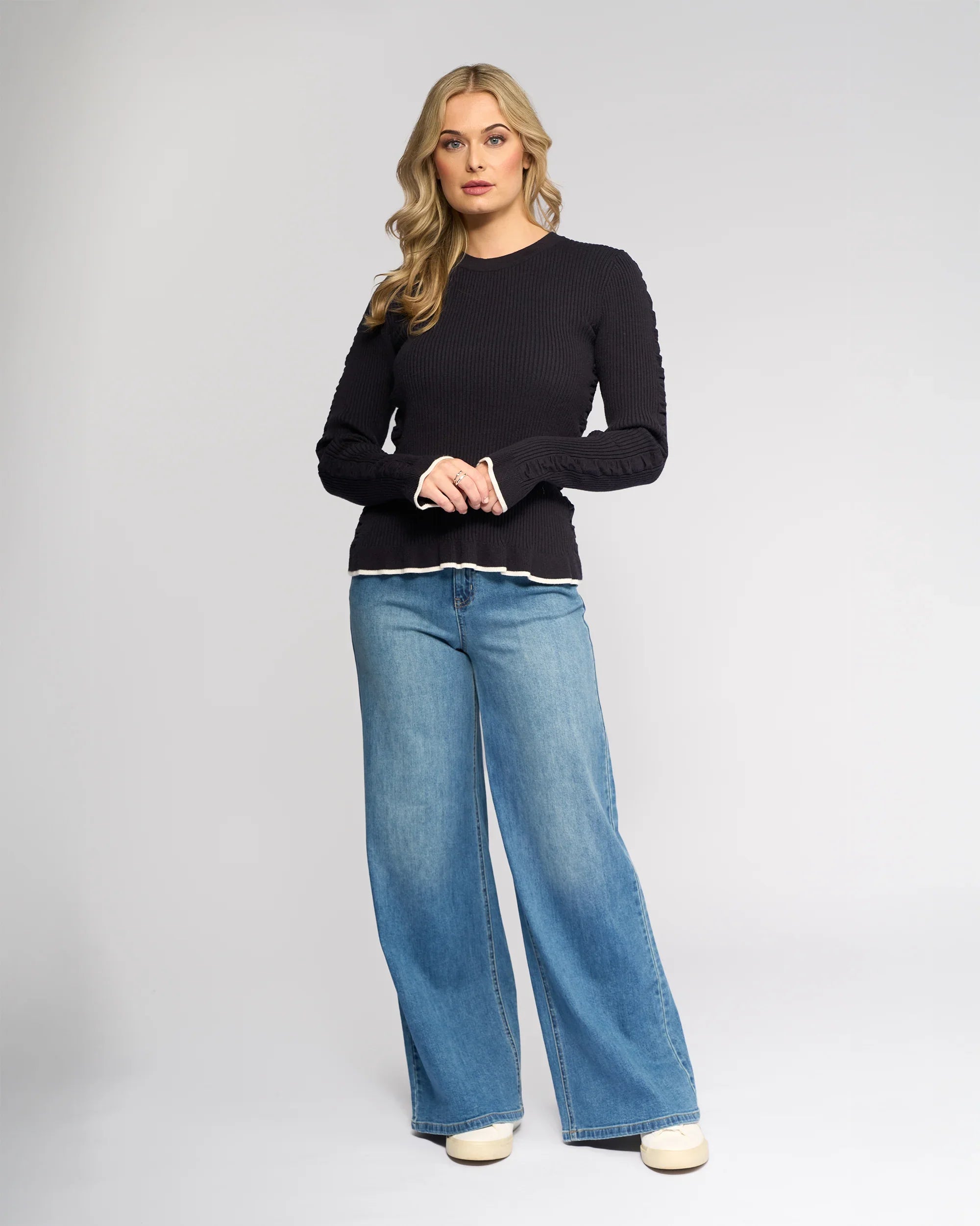 A woman with long blonde hair wears the Knewe Label Luxe Sweater in Ink/Winter White and wide-leg blue jeans, standing facing the camera against a plain light gray background.