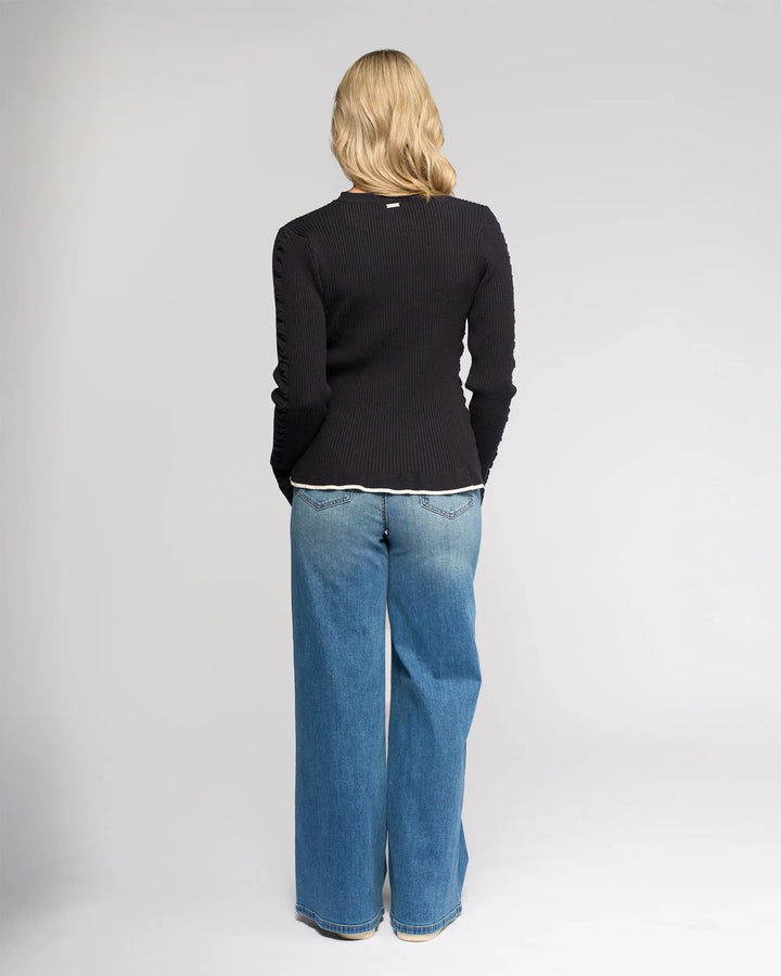 A person with blonde hair, wearing the Knewe Label Luxe Sweater in Ink/Winter White and wide-leg blue jeans, stands facing away from the camera against a plain light background.