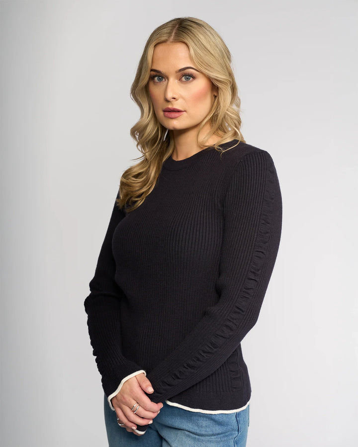 A woman with long blonde hair wears the Knewe Label Luxe Sweater in Ink/Winter White and blue jeans, standing against a plain light gray background and looking at the camera with a serious expression.