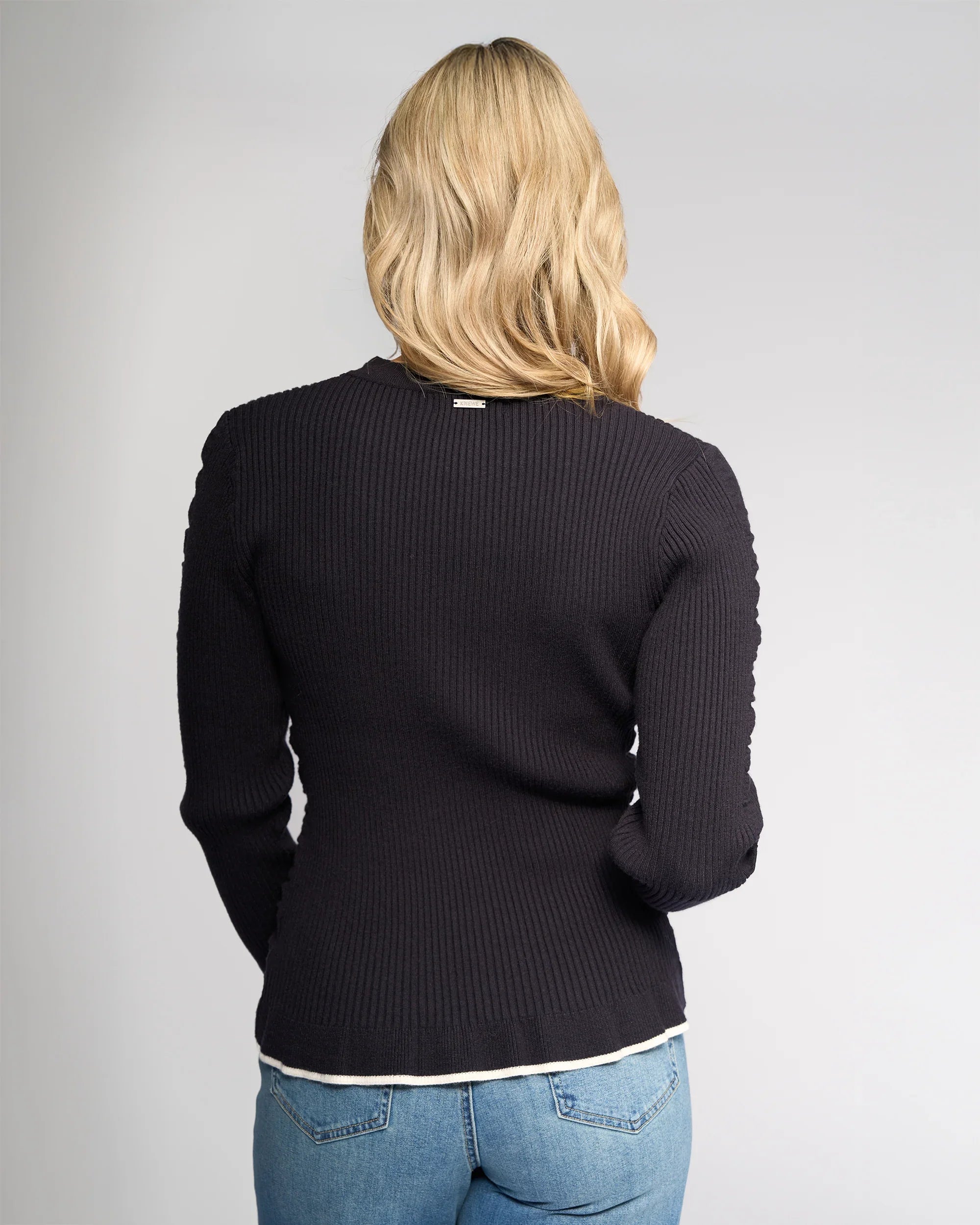A person with blonde, wavy hair is shown from the back, wearing the Luxe Sweater in Ink/Winter White by Knewe Label and blue jeans against a plain light gray background.