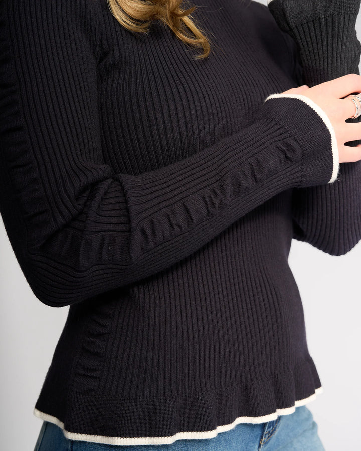 A person is shown from the torso up wearing the Knewe Label Luxe Sweater in Ink/Winter White—a slim-fit, ribbed knit sweater with white trim at the cuffs and hem—paired with blue jeans.