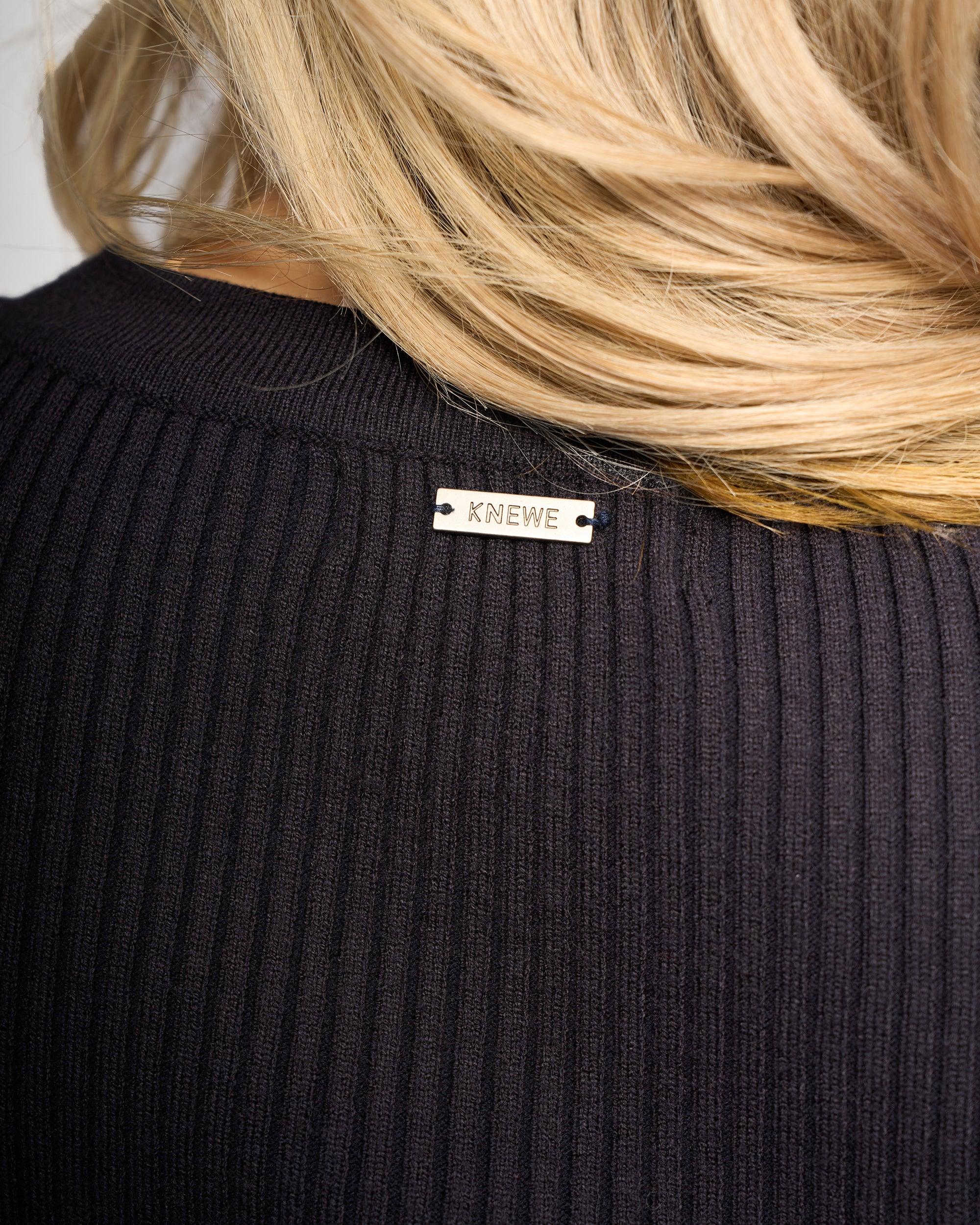 Close-up of the back of a person with blonde hair wearing the Luxe Sweater - Ink/Winter White by Knewe Label, featuring a ribbed knit texture and a small metallic brand label at the neckline.