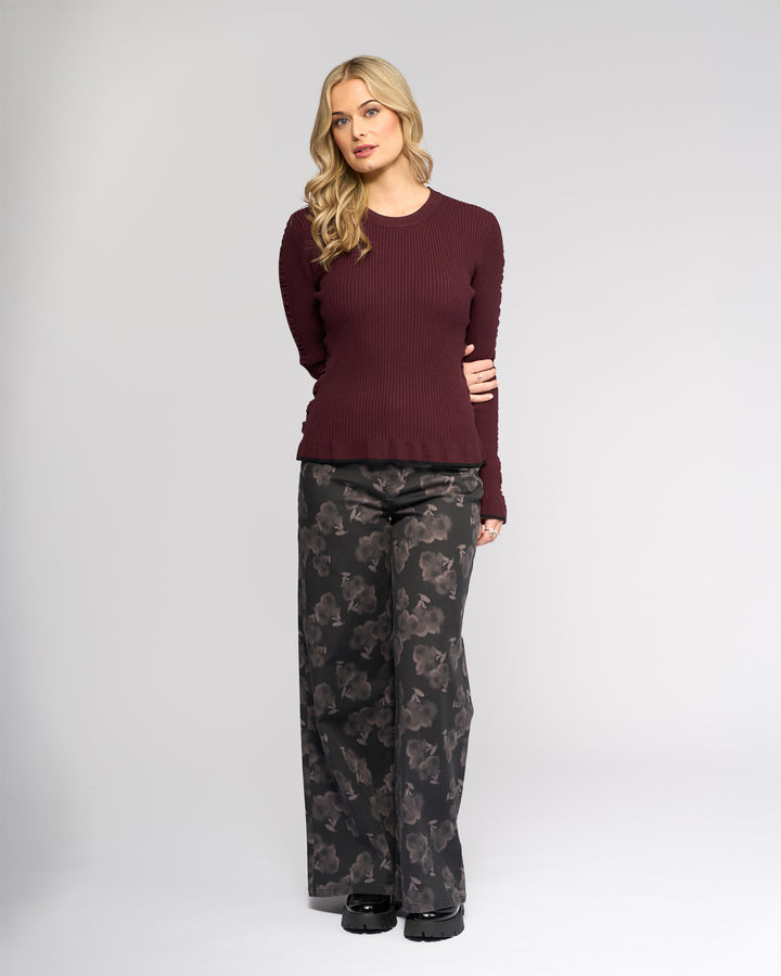Against a light gray background, a woman with long blonde hair wears the Knewe Label Luxe Sweater in Raisin/Black and loose, dark floral-patterned pants with black shoes. She poses with a neutral expression.