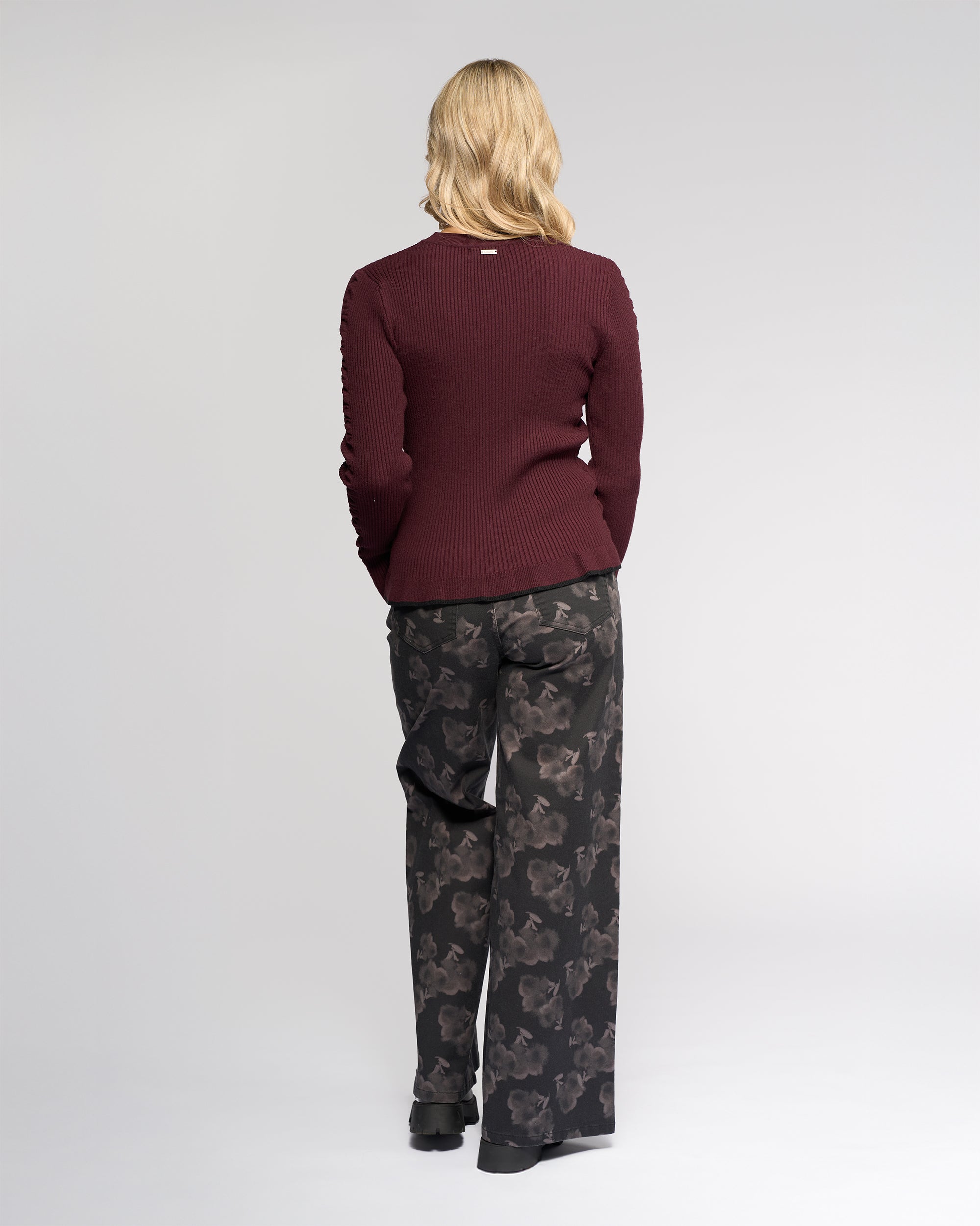 A person with shoulder-length blonde hair faces away, wearing the Knewe Label Luxe Sweater in Raisin/Black and loose black floral pants, set against a plain light background.