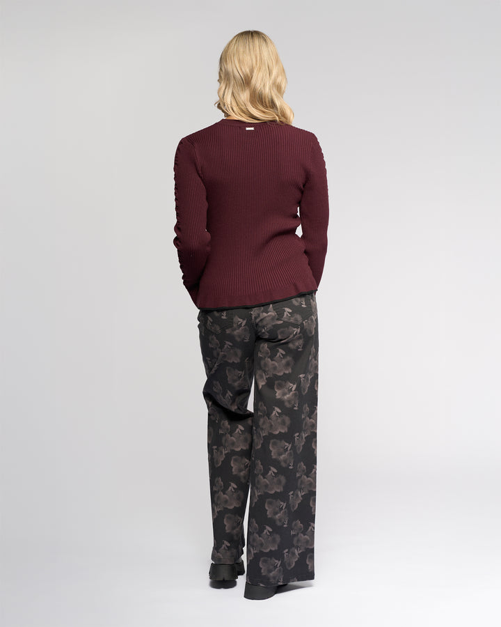A person with shoulder-length blonde hair faces away, wearing the Knewe Label Luxe Sweater in Raisin/Black and loose black floral pants, set against a plain light background.
