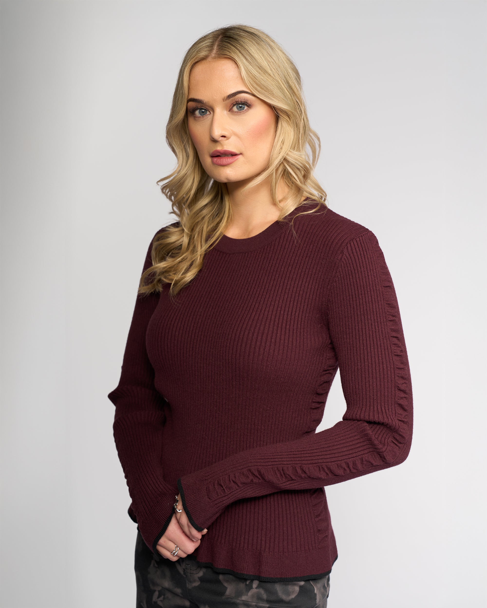 A woman with long blonde hair models the Knewe Label Luxe Sweater in Raisin/Black and dark patterned pants against a light gray background, gazing slightly to the side with a neutral expression.
