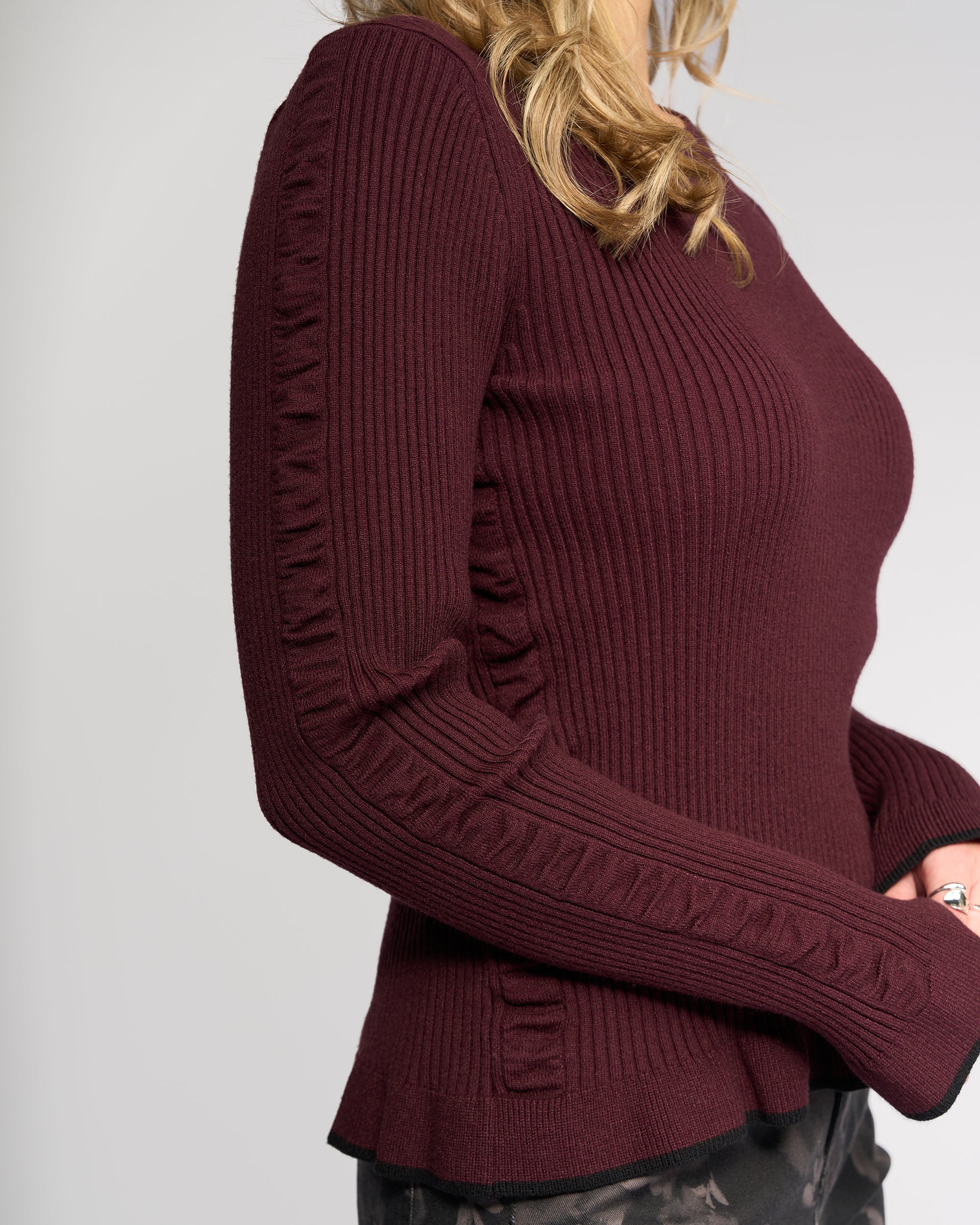 A woman with wavy blonde hair wears the Knewe Label Luxe Sweater in Raisin/Black, a slim-fit ribbed knit with ruffle accents and textured side panels. Only her upper body is shown.