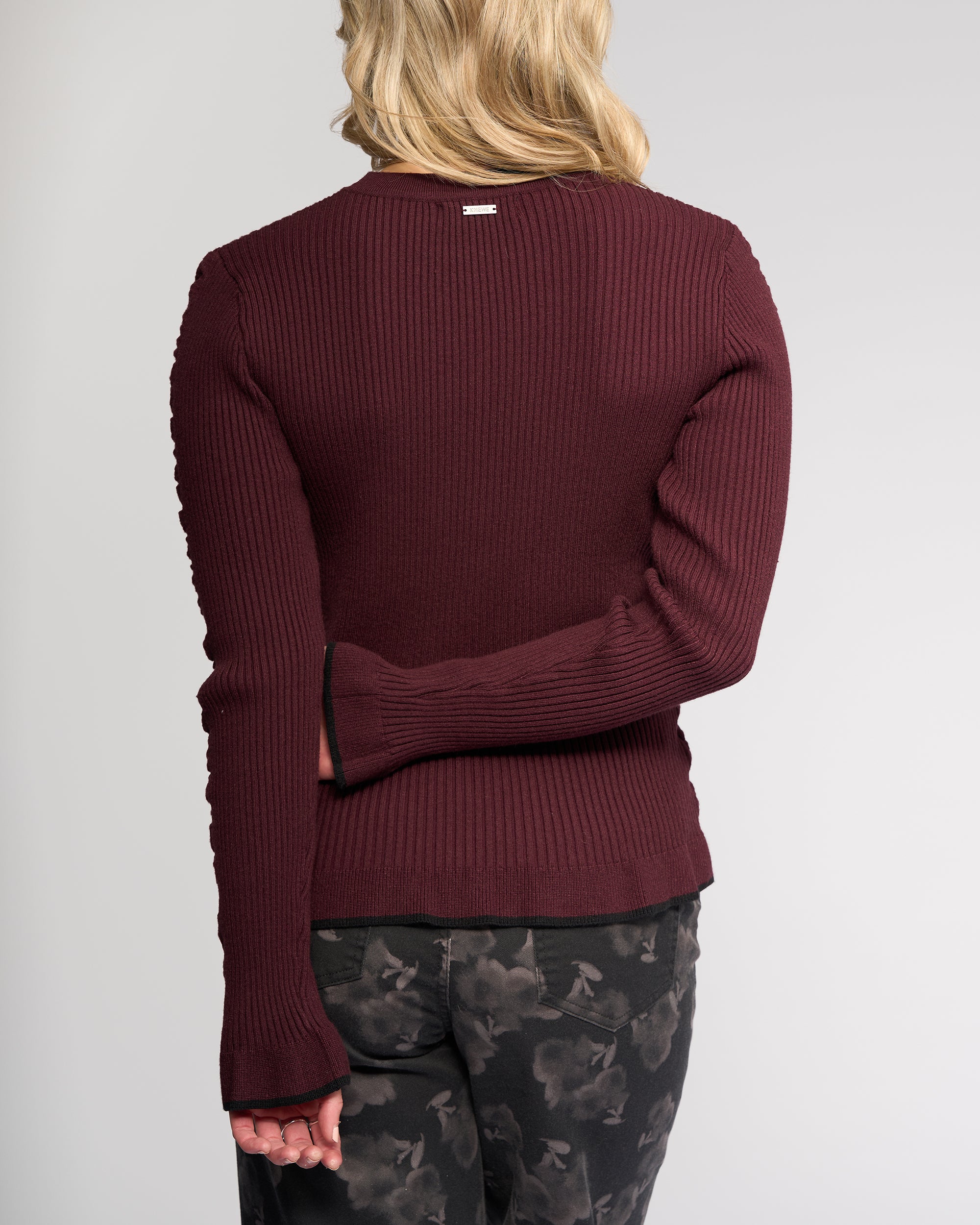 A person with shoulder-length blonde hair is seen from behind, wearing the Knewe Label Luxe Sweater in Raisin/Black with dark patterned pants. One arm is bent behind their back while the other hangs by their side.