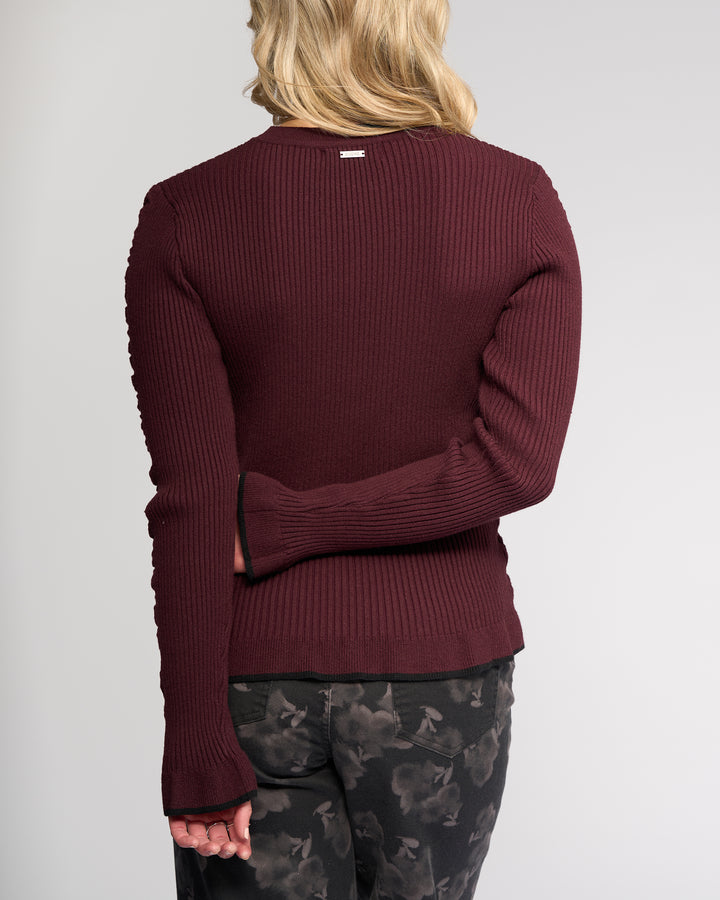 A person with shoulder-length blonde hair is seen from behind, wearing the Knewe Label Luxe Sweater in Raisin/Black with dark patterned pants. One arm is bent behind their back while the other hangs by their side.