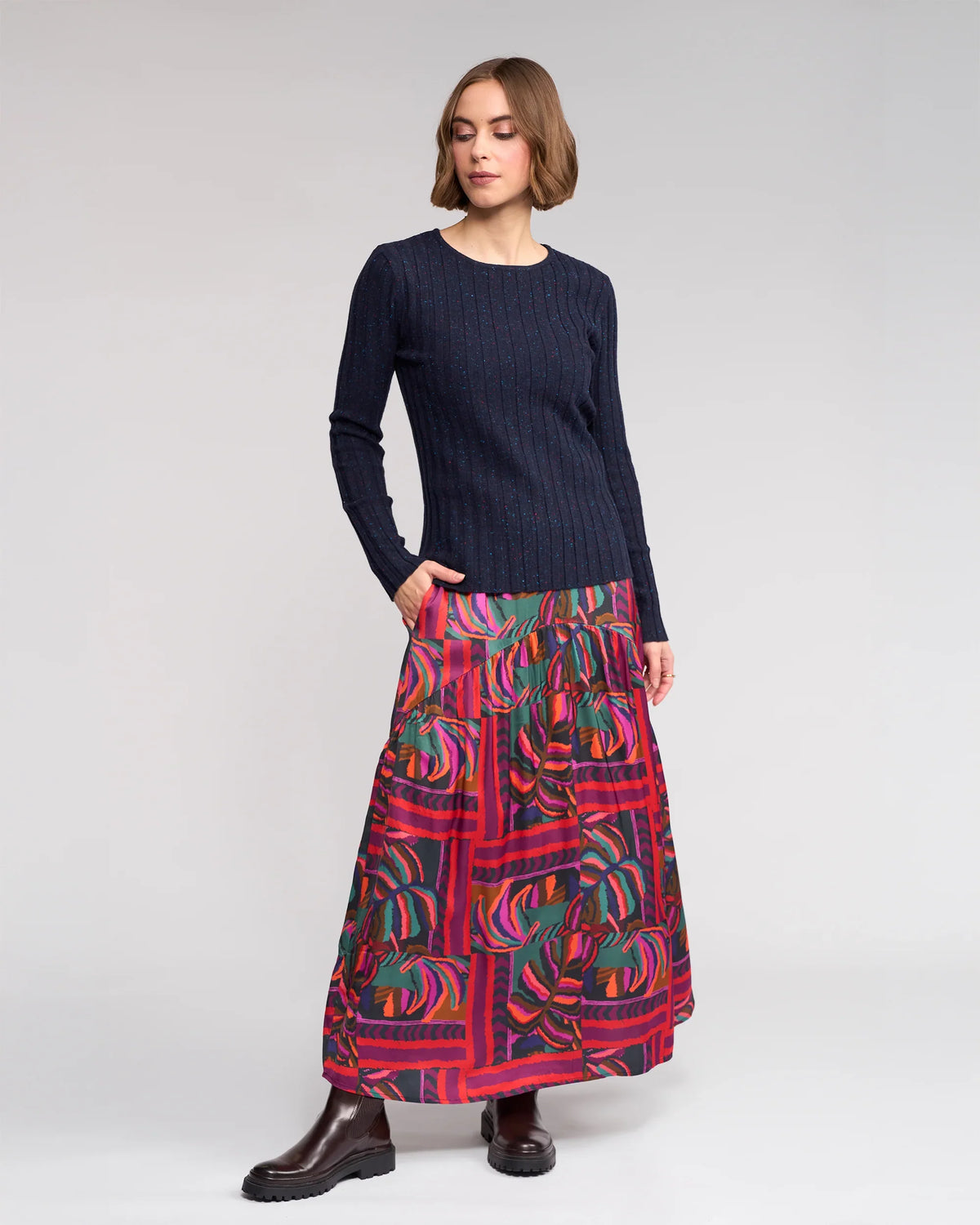 A woman stands against a light gray background wearing the Knewe Label Felix Sweater in Navy Fleck, paired with a long colorful abstract floral skirt in red, green, and purple tones, and dark brown ankle boots.