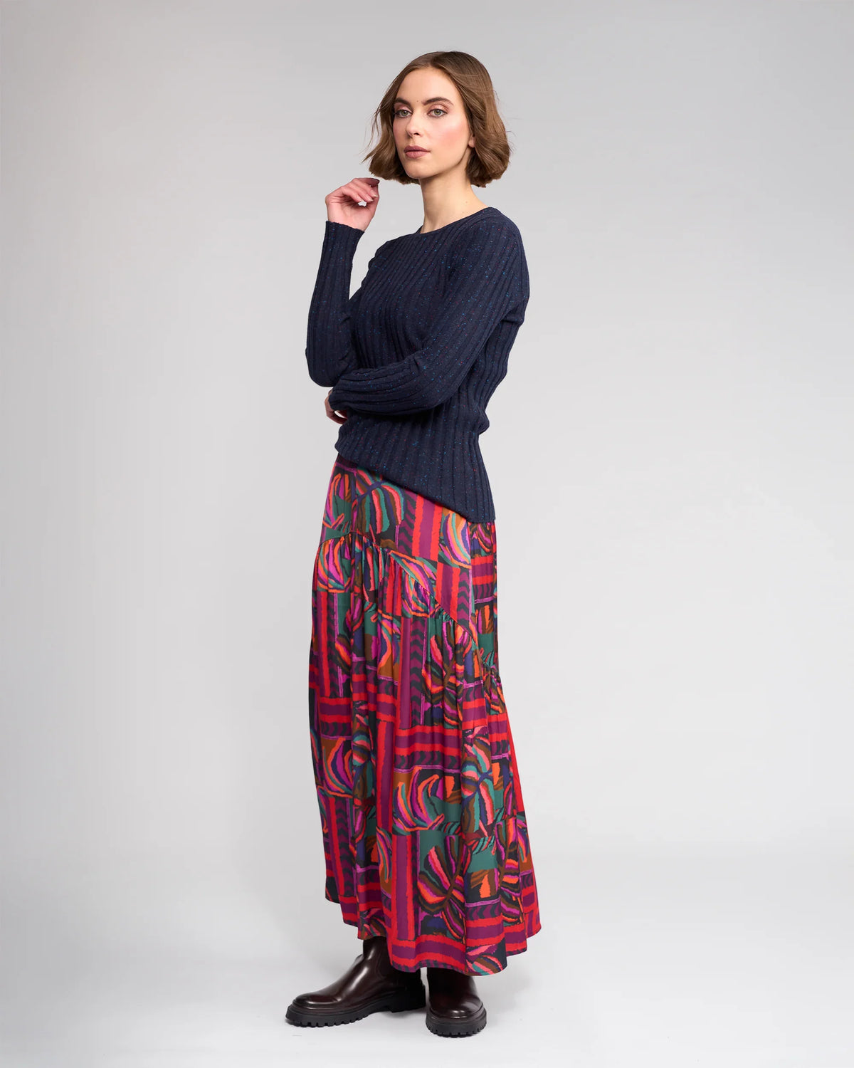 A woman with short brown hair and a thoughtful expression stands in profile against a plain background, wearing the Knewe Label Felix Sweater in Navy Fleck, paired with a bold abstract long skirt and dark ankle boots.