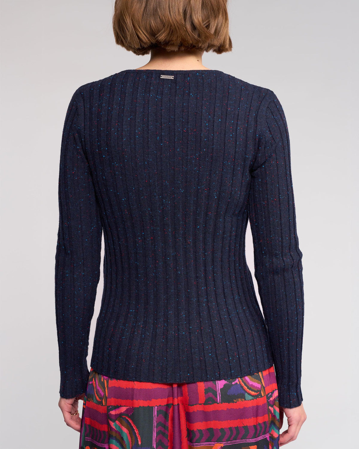 A person with short brown hair is seen from behind, wearing the Knewe Label Felix Sweater in Navy Fleck and a colorful patterned skirt, set against a plain background.