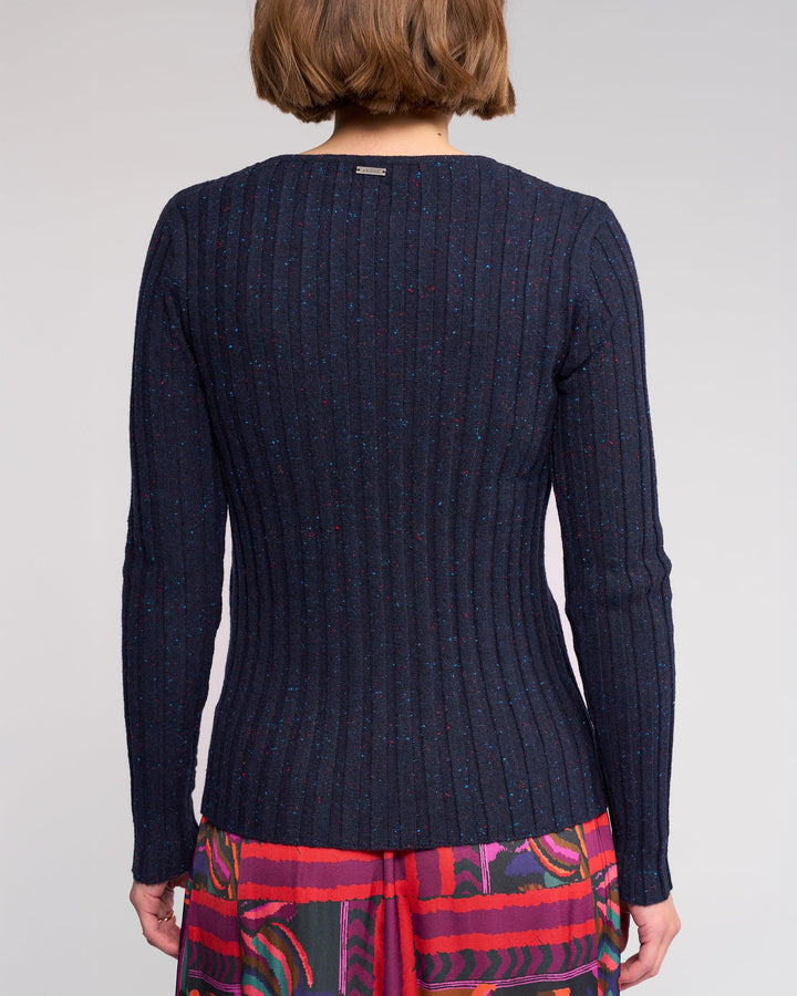 A person with short brown hair is seen from behind, wearing the Knewe Label Felix Sweater in Navy Fleck and a colorful patterned skirt, set against a plain background.