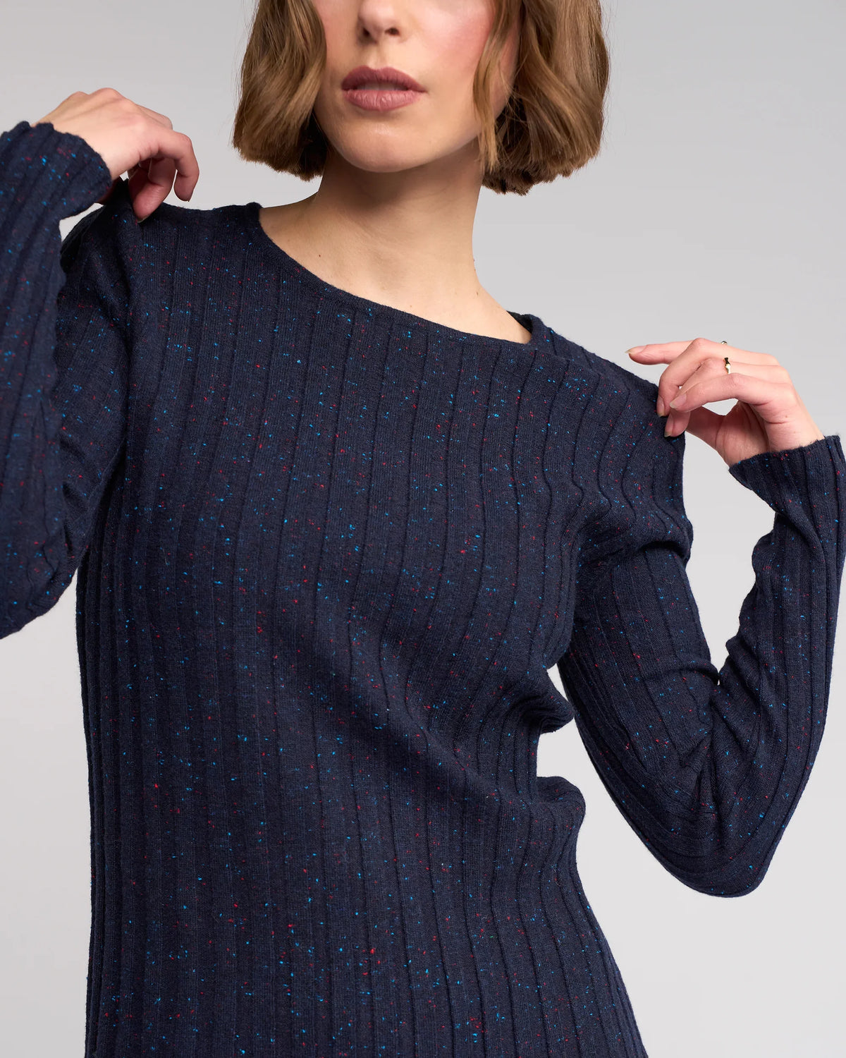 A woman with short brown hair models the Knewe Label Felix Sweater in Navy Fleck—a slim fit ribbed knit with colorful specks—posing with one arm bent and her hand near her chin, looking slightly to the side.