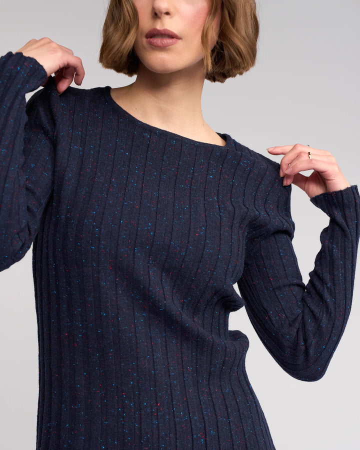 A woman with short brown hair models the Knewe Label Felix Sweater in Navy Fleck—a slim fit ribbed knit with colorful specks—posing with one arm bent and her hand near her chin, looking slightly to the side.