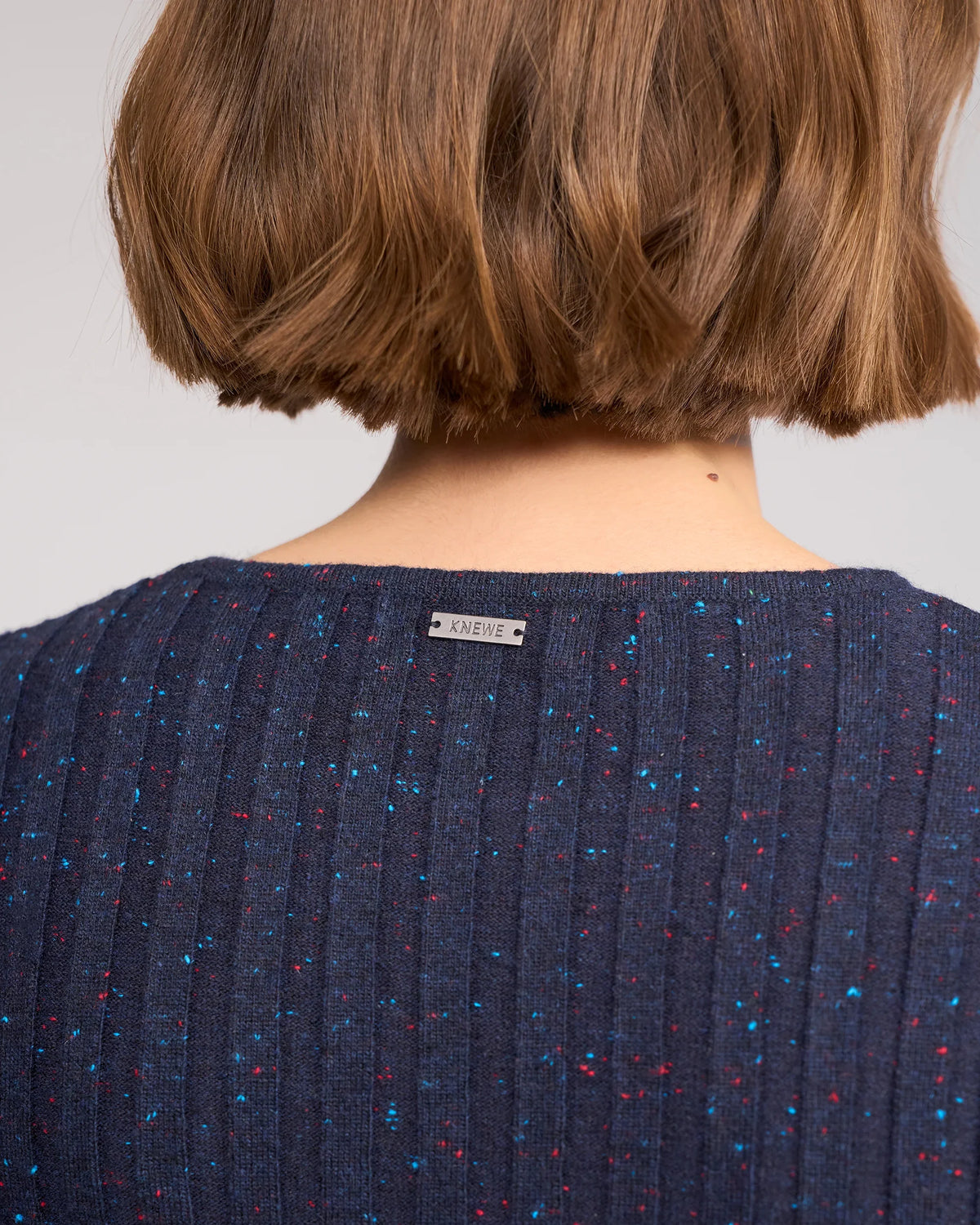 A close-up shows the back of a person with short brown hair wearing the Knewe Label Felix Sweater in Navy Fleck, a slim-fit ribbed knit featuring red and blue specks and a small KNEWE tag at the neckline.