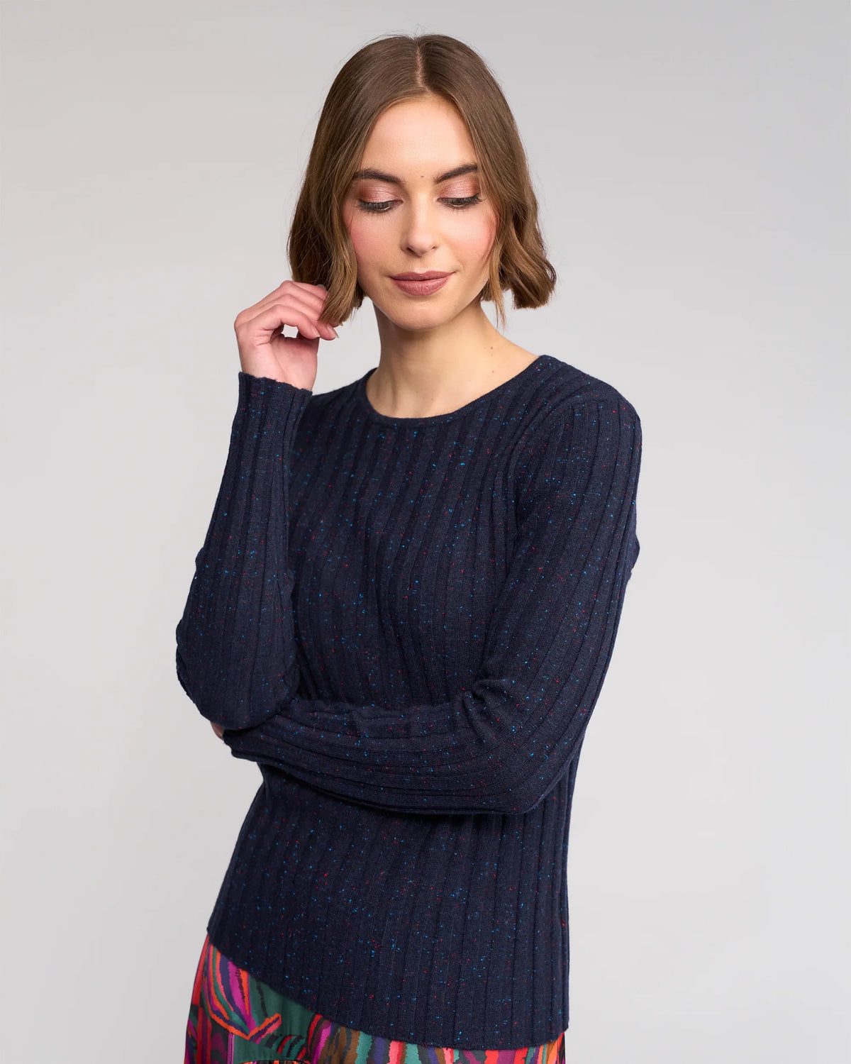A woman with light brown hair wears the Felix Sweater - Navy Fleck by Knewe Label, paired with a colorful skirt against a light gray background. She looks down with a gentle smile, her hand near her face.