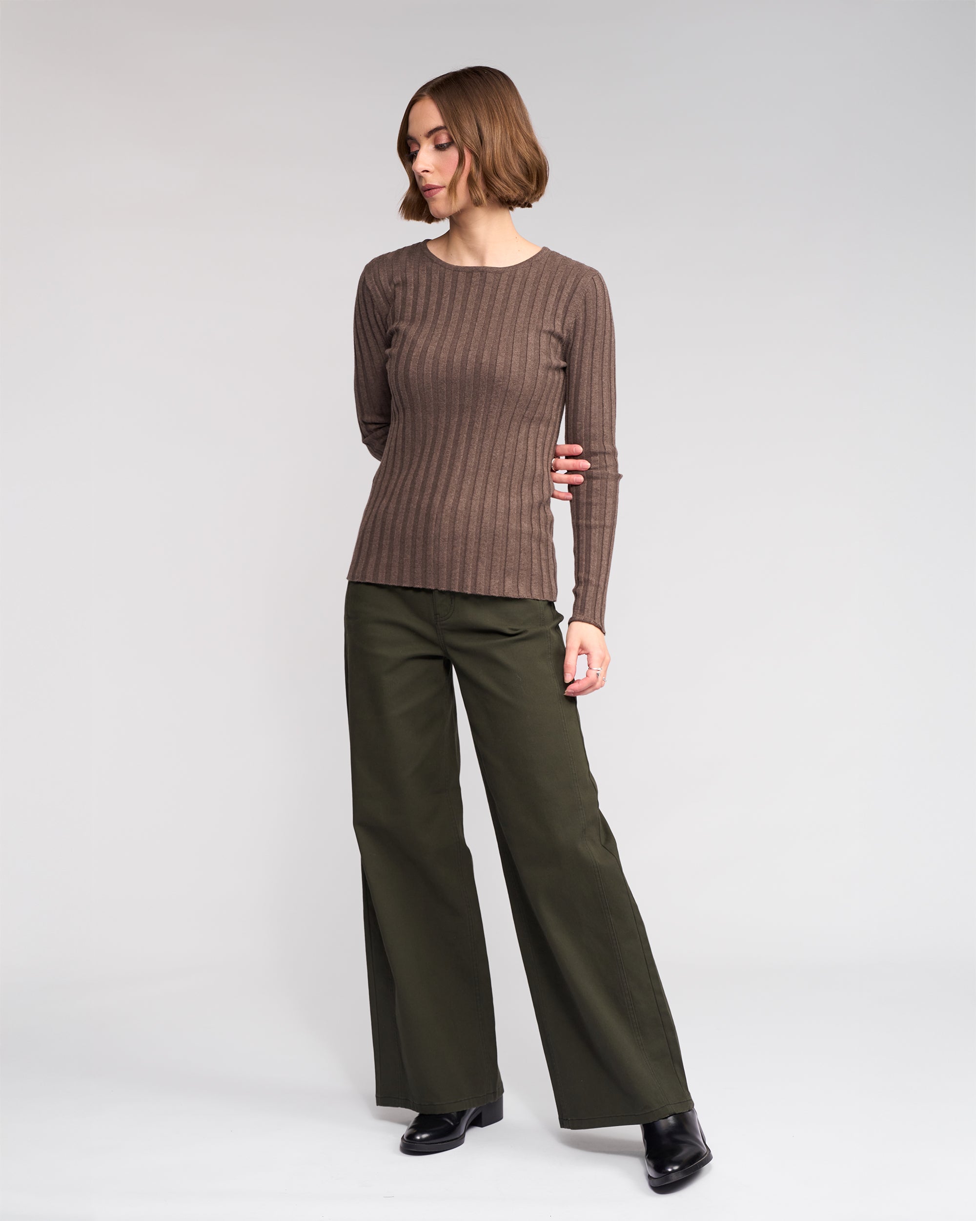 A woman with short brown hair wears the Knewe Label Felix Sweater in Truffle with wide-leg olive green pants and black shoes, standing against a plain light background.