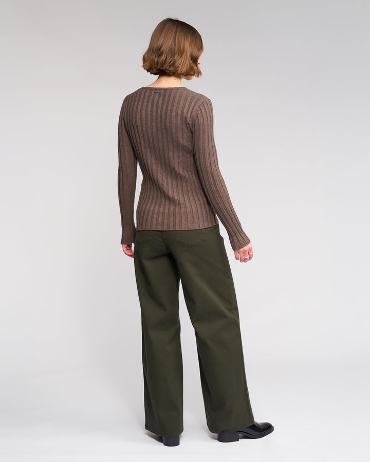 A person with short brown hair faces away, wearing the Knewe Label Felix Sweater in Truffle, dark green wide-leg pants, and black shoes against a plain light background.